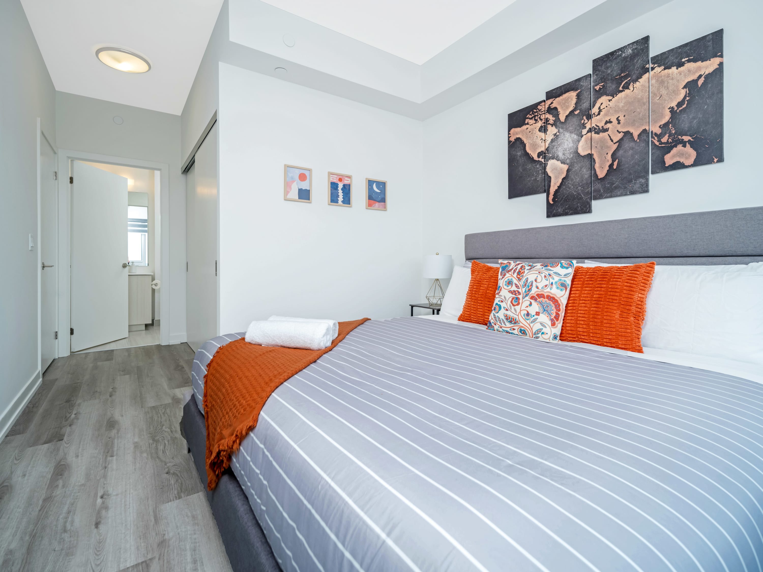 Cozy bedroom featuring a gray upholstered bed with a striped duvet, orange throw blanket, and decorative pillows, flanked by a nightstand with a lamp, and a large world map artwork above the headboard.