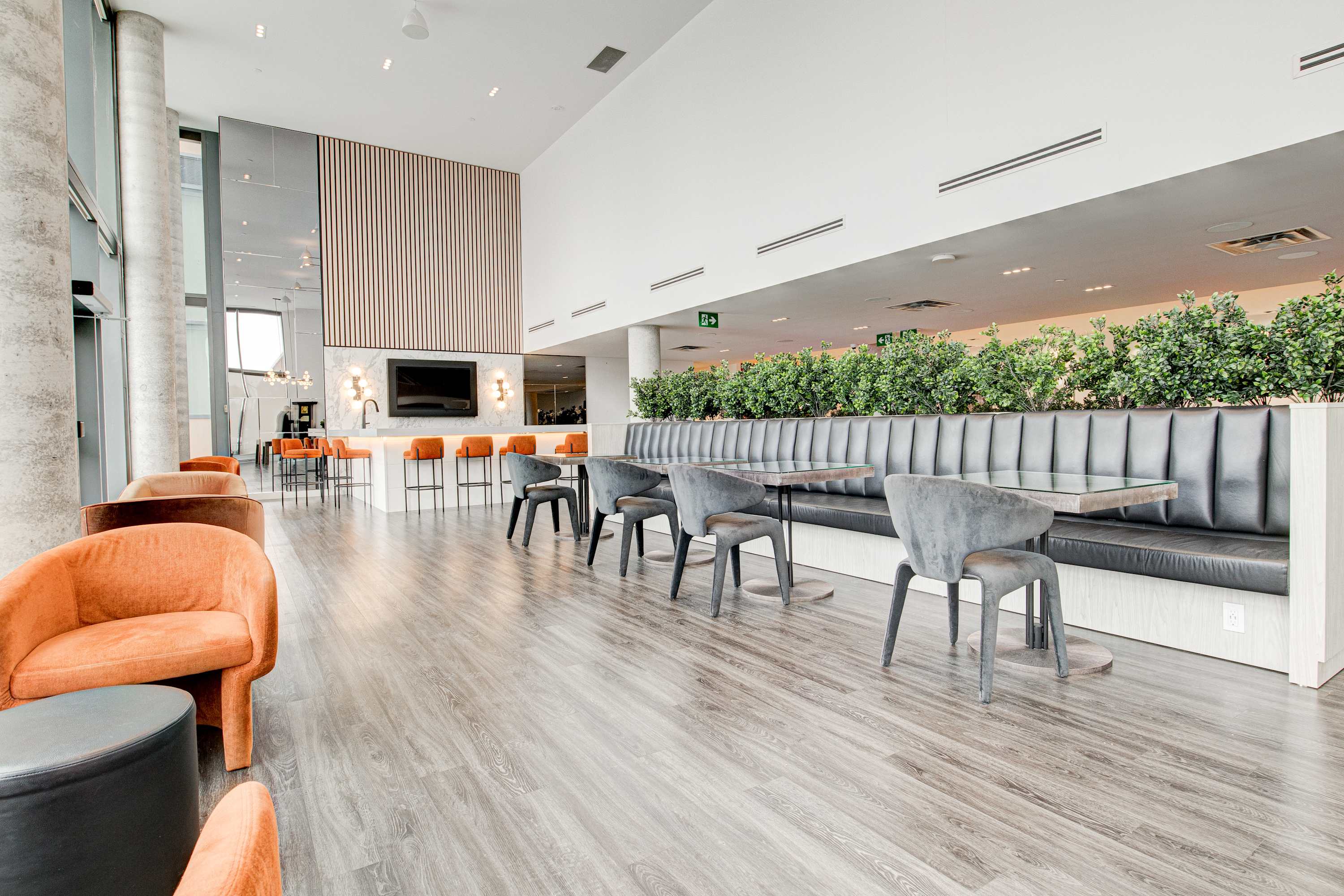 A spacious and modern common lounge area features long booth seating with dark upholstery, individual tables with grey chairs, and vibrant orange armchairs. A bar area with high stools and a large TV is visible in the background, with plants along the wall.