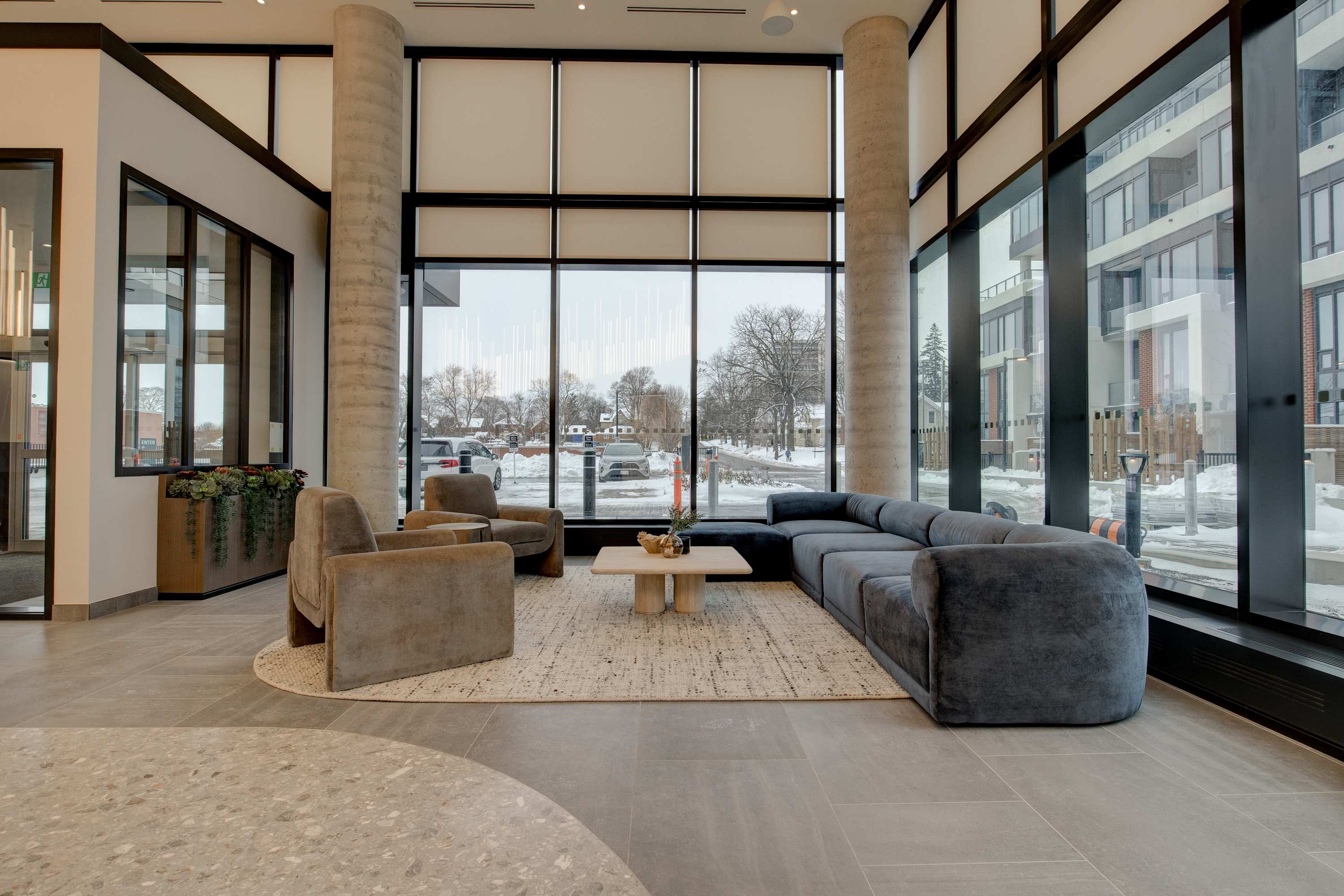 A spacious, modern lounge area features large floor-to-ceiling windows with city views, a dark grey L-shaped sectional sofa, and two light grey armchairs. Concrete pillars frame the space, which is set on light grey tiled flooring with a textured area rug.