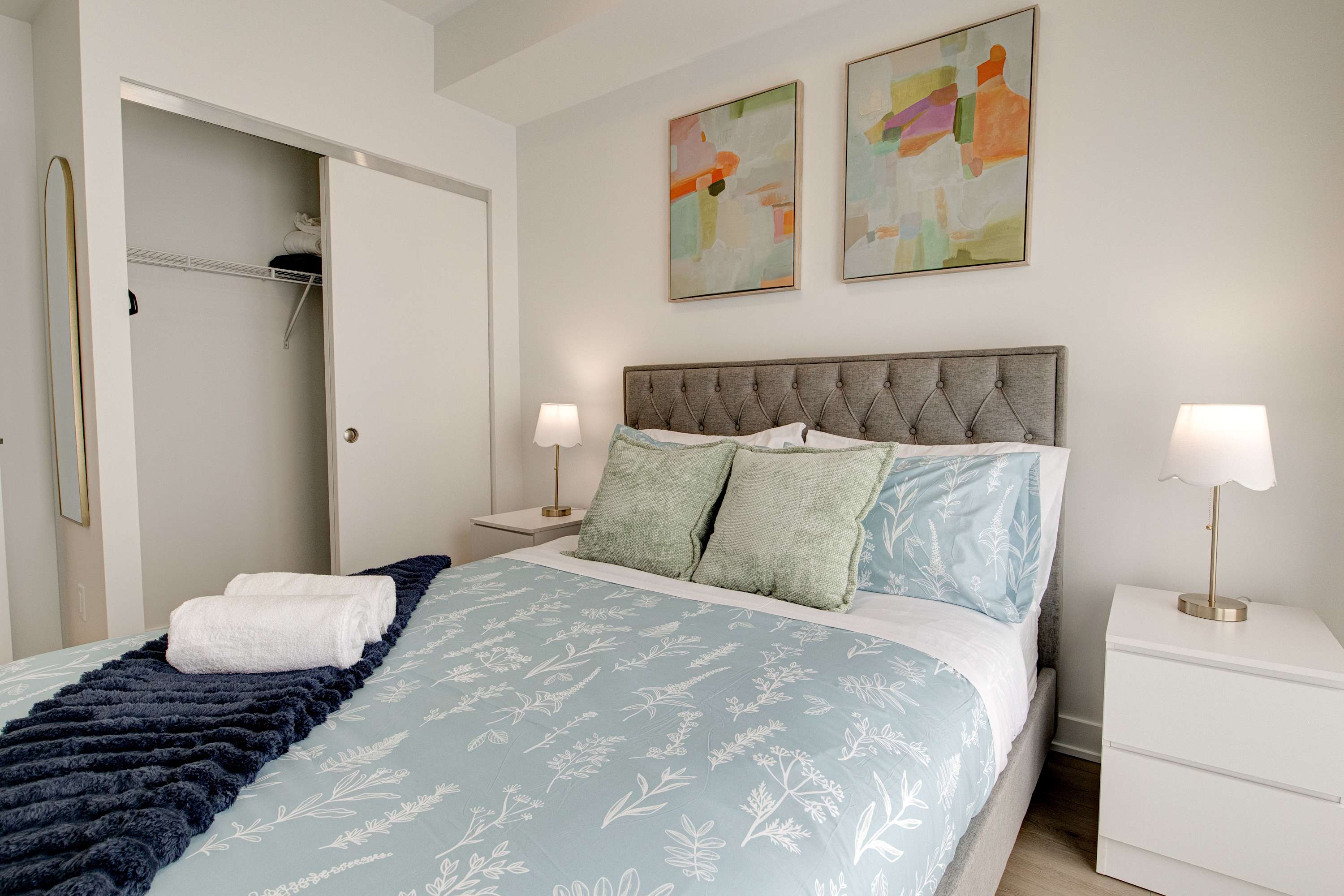 A cozy bedroom features a queen-sized bed with a tufted grey headboard, light blue and green bedding, and two abstract art pieces above. A sliding closet door is open to the left, revealing shelving.