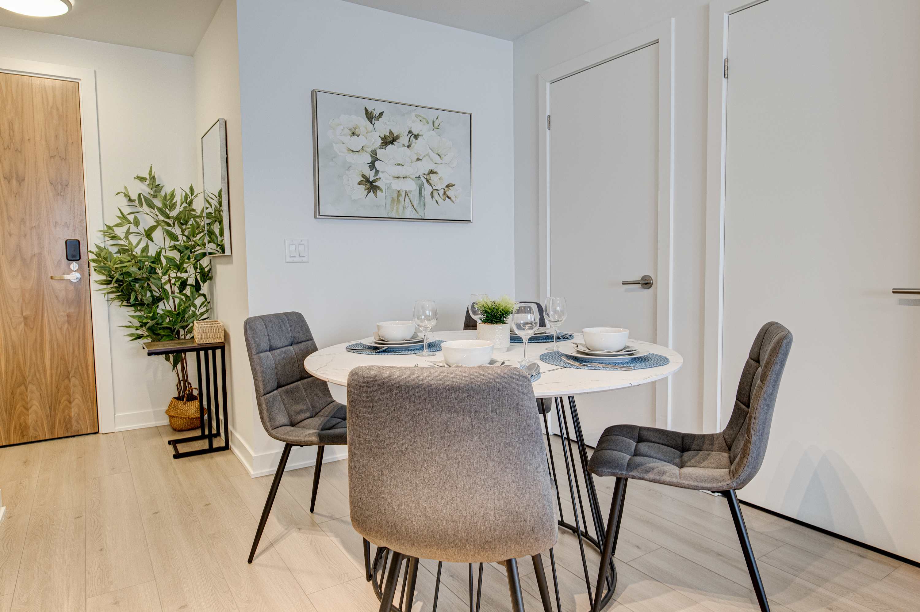 Dining area with a round white table and four grey chairs, positioned near the entrance with a wooden door, a small console table with greenery, and floral artwork on the wall.