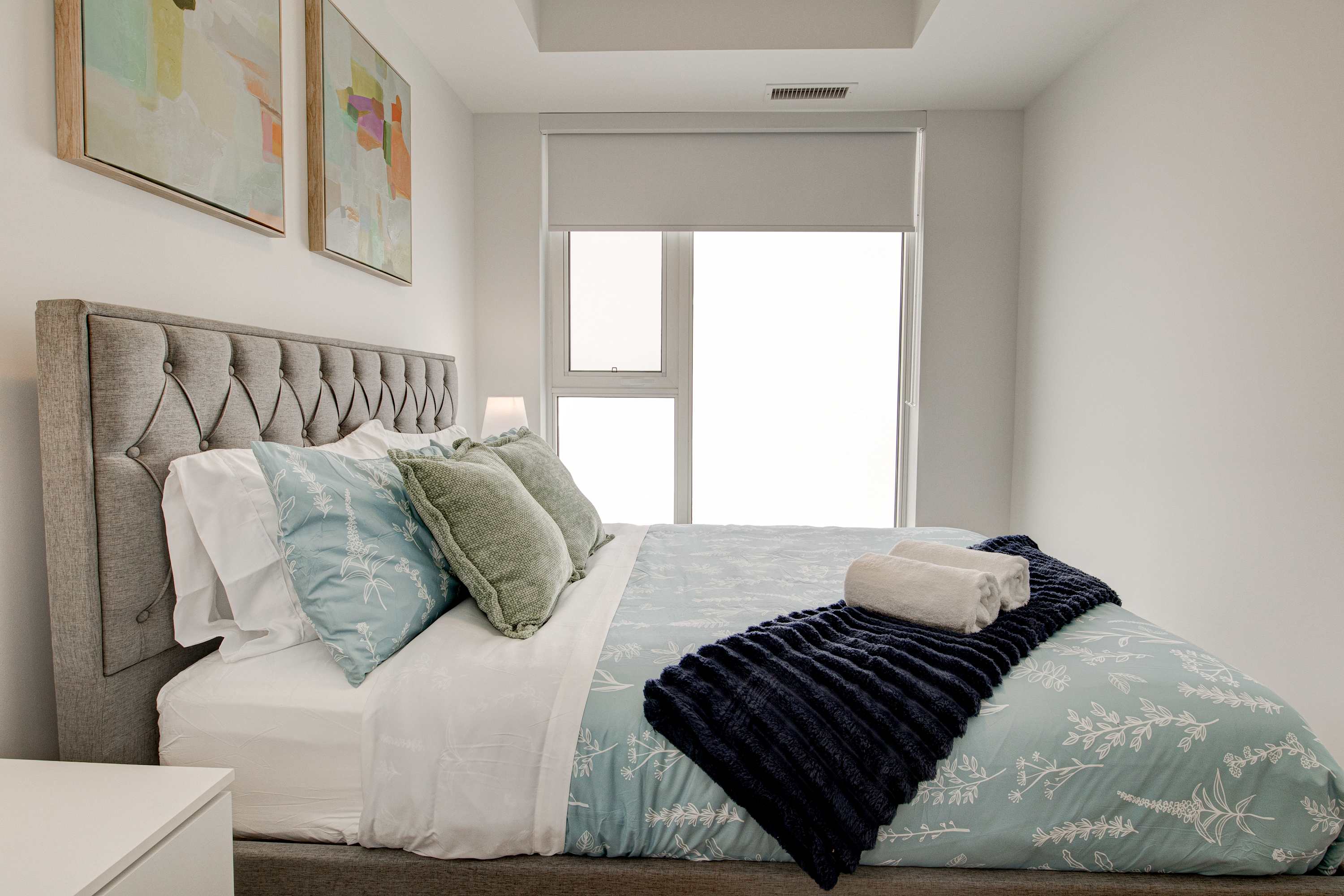 The bright bedroom features a queen bed with a grey tufted headboard and light blue bedding, accented by a dark blue throw blanket and white towels. Large windows with room-darkening shades offer natural light and a view of the outside.