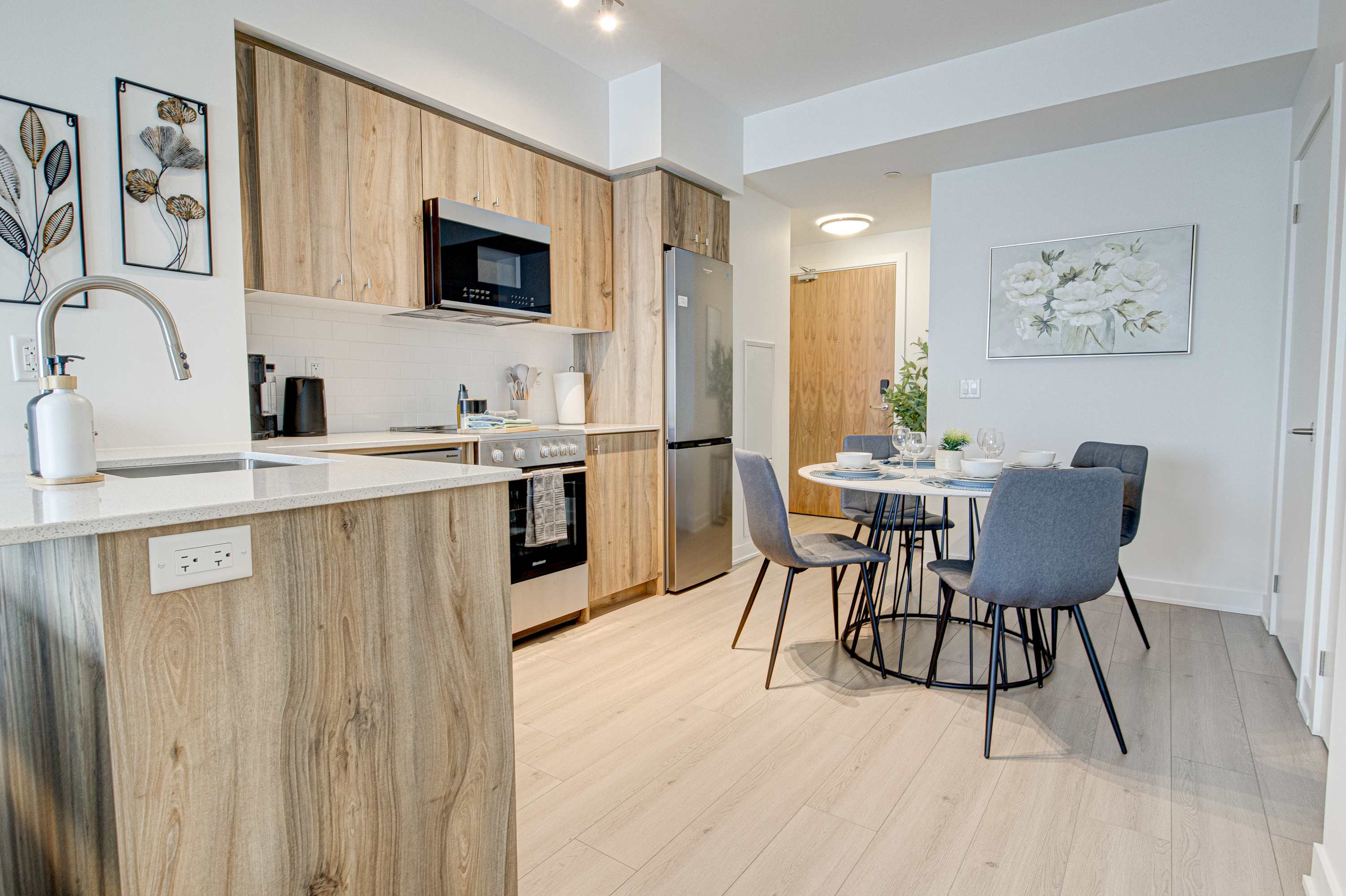 Open-concept kitchen and dining area featuring light wood cabinetry, stainless steel appliances, a white subway tile backsplash, and a round dining table with grey chairs.
