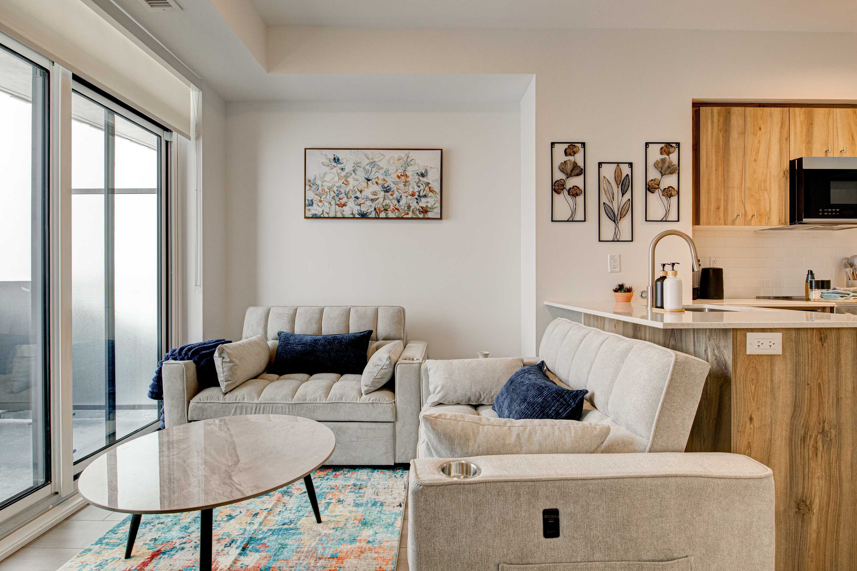 Bright living room with two light-colored sofas, one featuring a cup holder, a round glass coffee table on a colorful rug, and an open view towards the modern kitchen area.