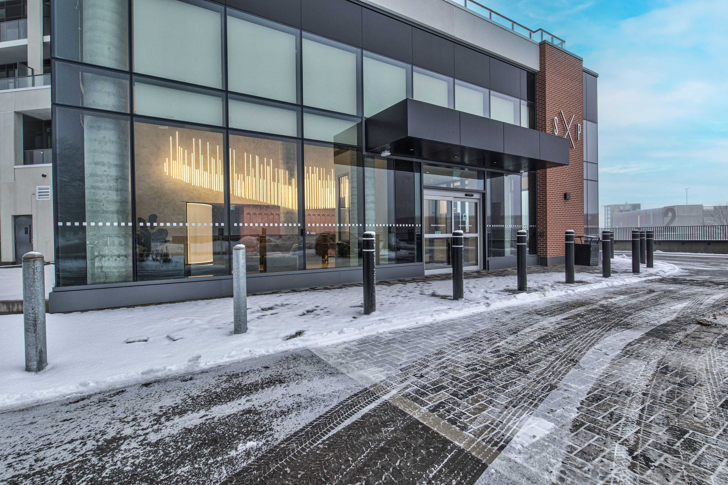 The modern building entrance features large glass windows, a black awning, and automatic doors, with a snowy paved driveway leading up to it. The exterior combines dark paneling and brick accents, with bollards lining the path.