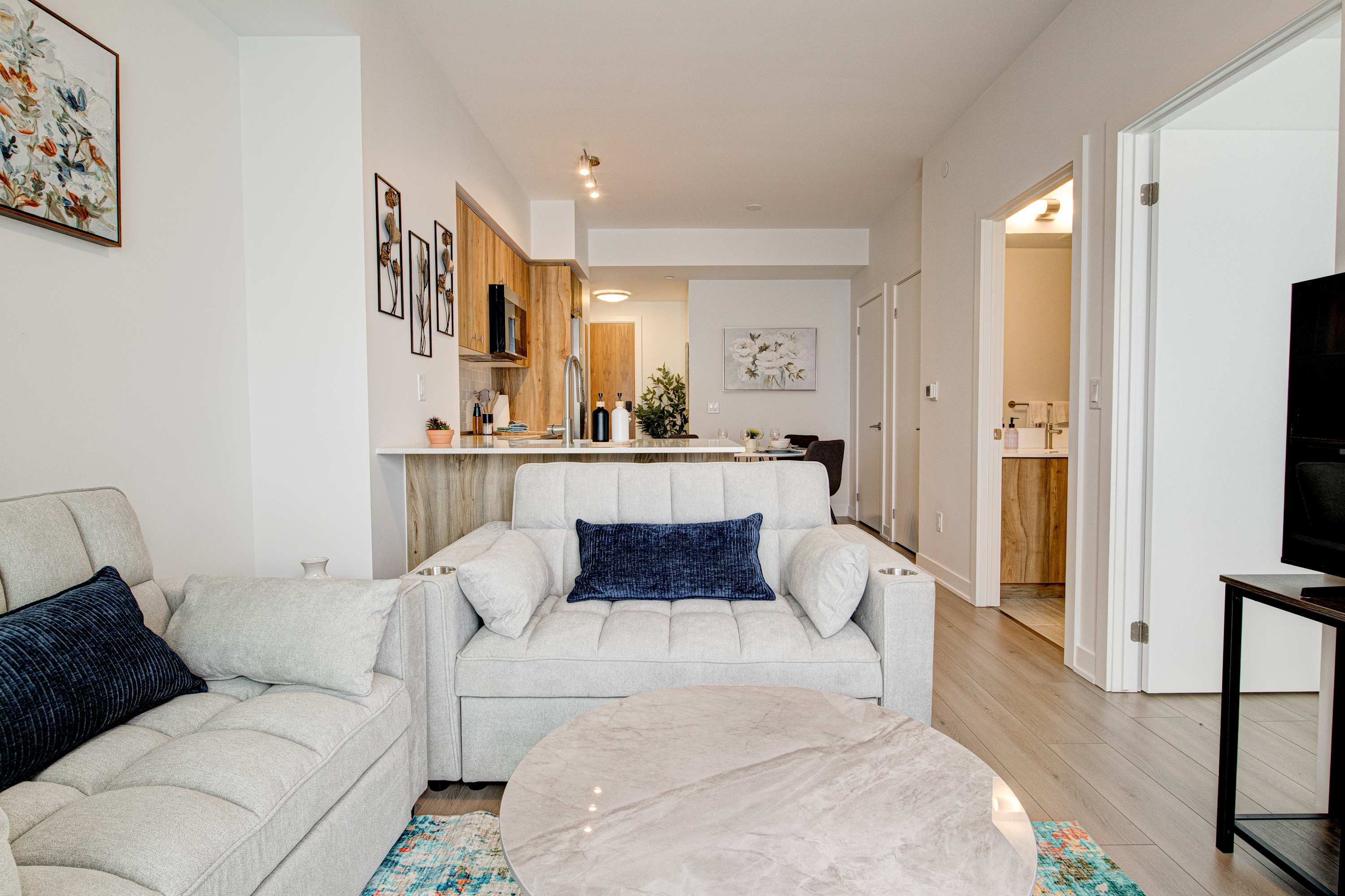 A modern open-concept living space features a light grey sofa with a dark blue throw pillow, a round marble coffee table, and light wood flooring. The kitchen with light wood cabinetry and a white countertop is visible in the background, along with a dining area.