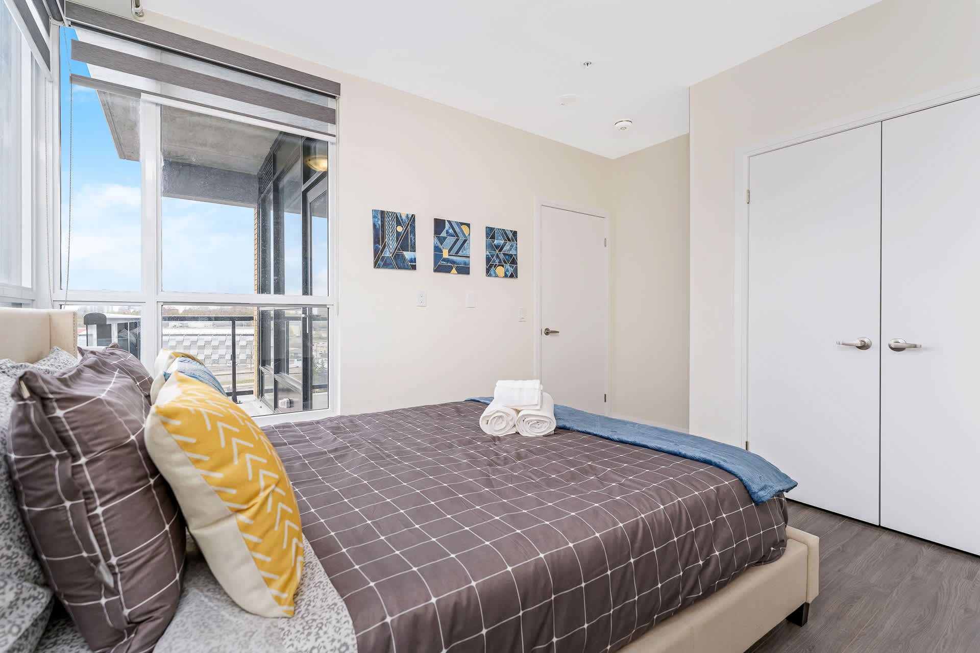 View of the bedroom showcasing a queen bed with patterned bedding and decorative pillows. The room features three abstract art pieces on the wall, a closet with white bi-fold doors, and large windows with blinds.