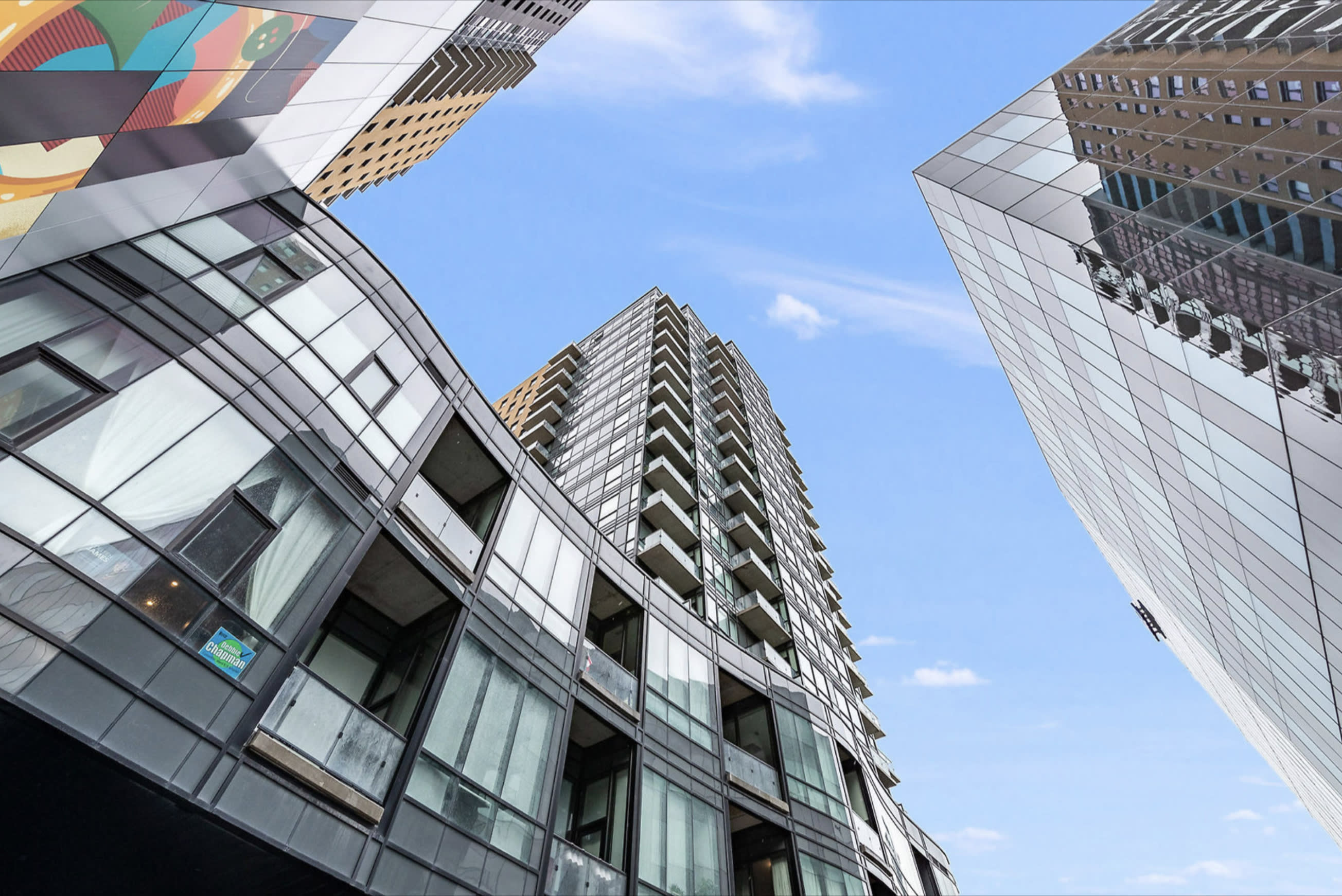 Dynamic upward view of contemporary high-rise buildings with reflective glass facades and multiple balconies, set against a clear blue sky with scattered clouds.