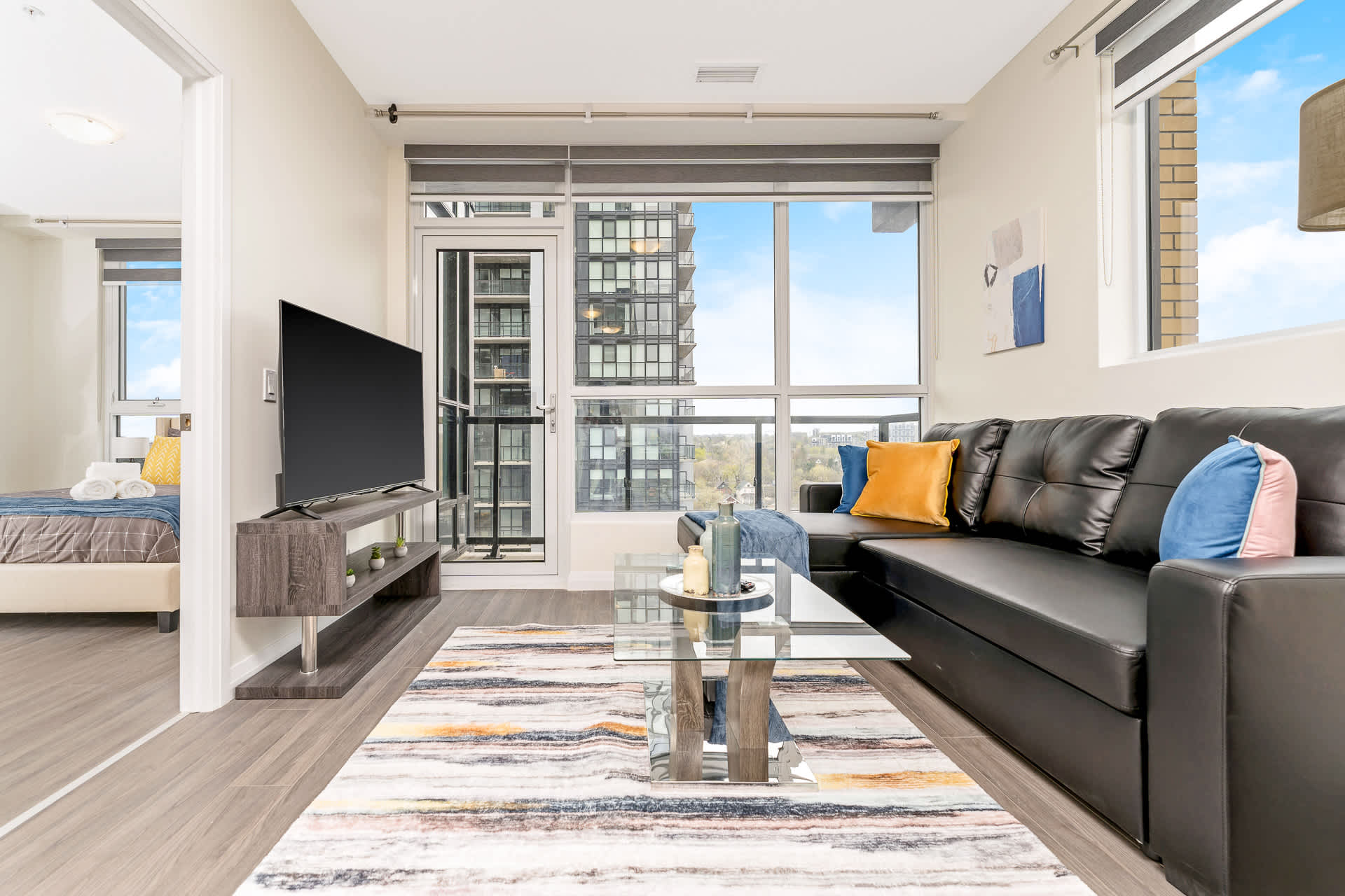 Modern living room featuring a black sectional sofa, glass coffee table, and a colorful abstract rug on dark wood-look flooring. Floor-to-ceiling windows offer city views and lead to a balcony, with a flat-screen TV on a dark wood stand.
