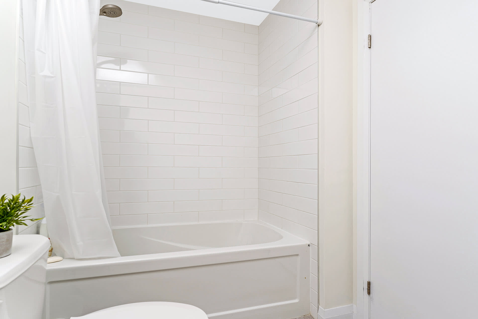 Bright bathroom with a white bathtub and shower combo, enclosed by a white shower curtain. The walls are tiled with white subway tiles, and a portion of the toilet is visible.