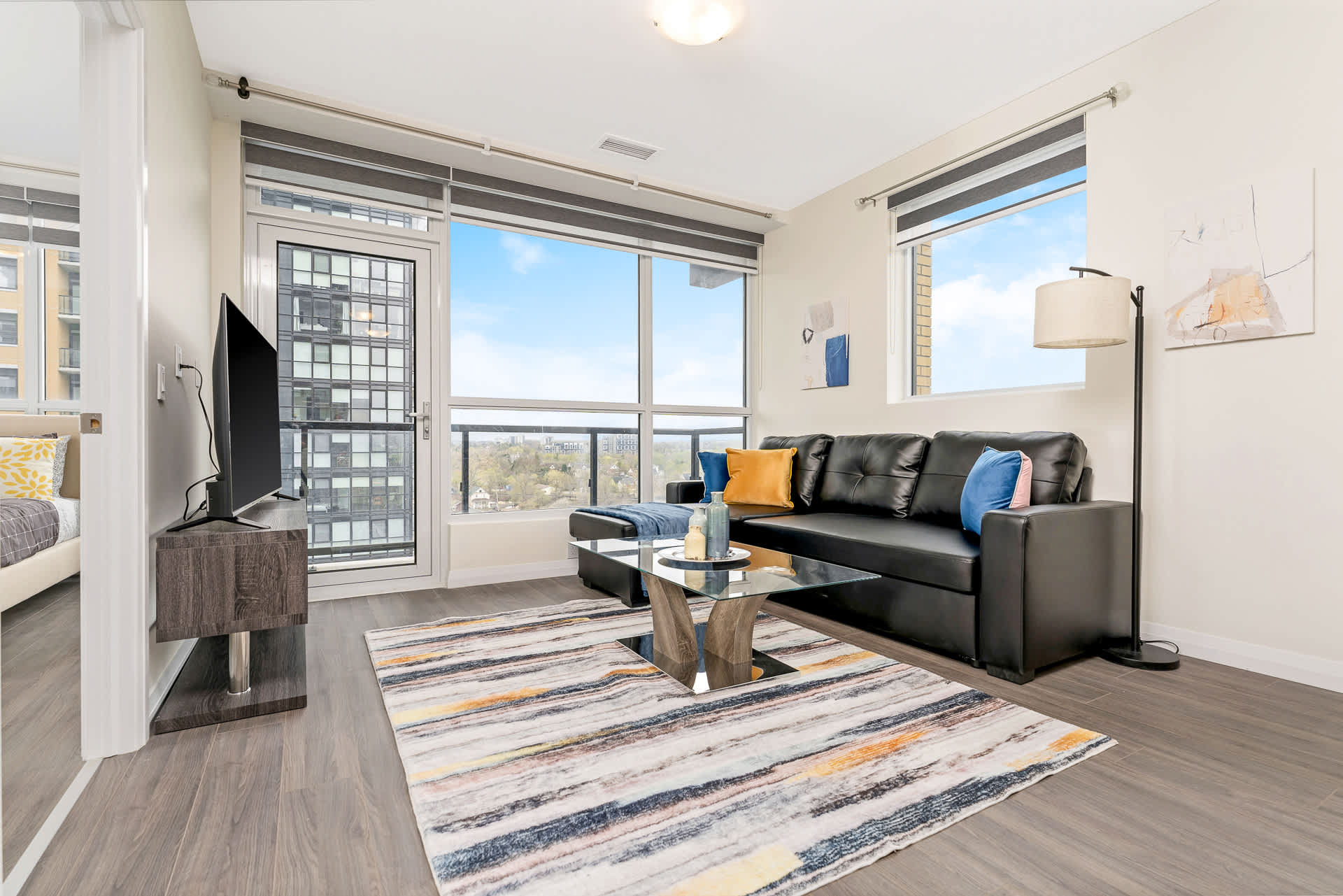 Bright living room featuring a black sectional sofa with colorful throw pillows, a glass coffee table, and a vibrant abstract rug. Large windows provide ample natural light and city views, with a floor lamp in the corner.