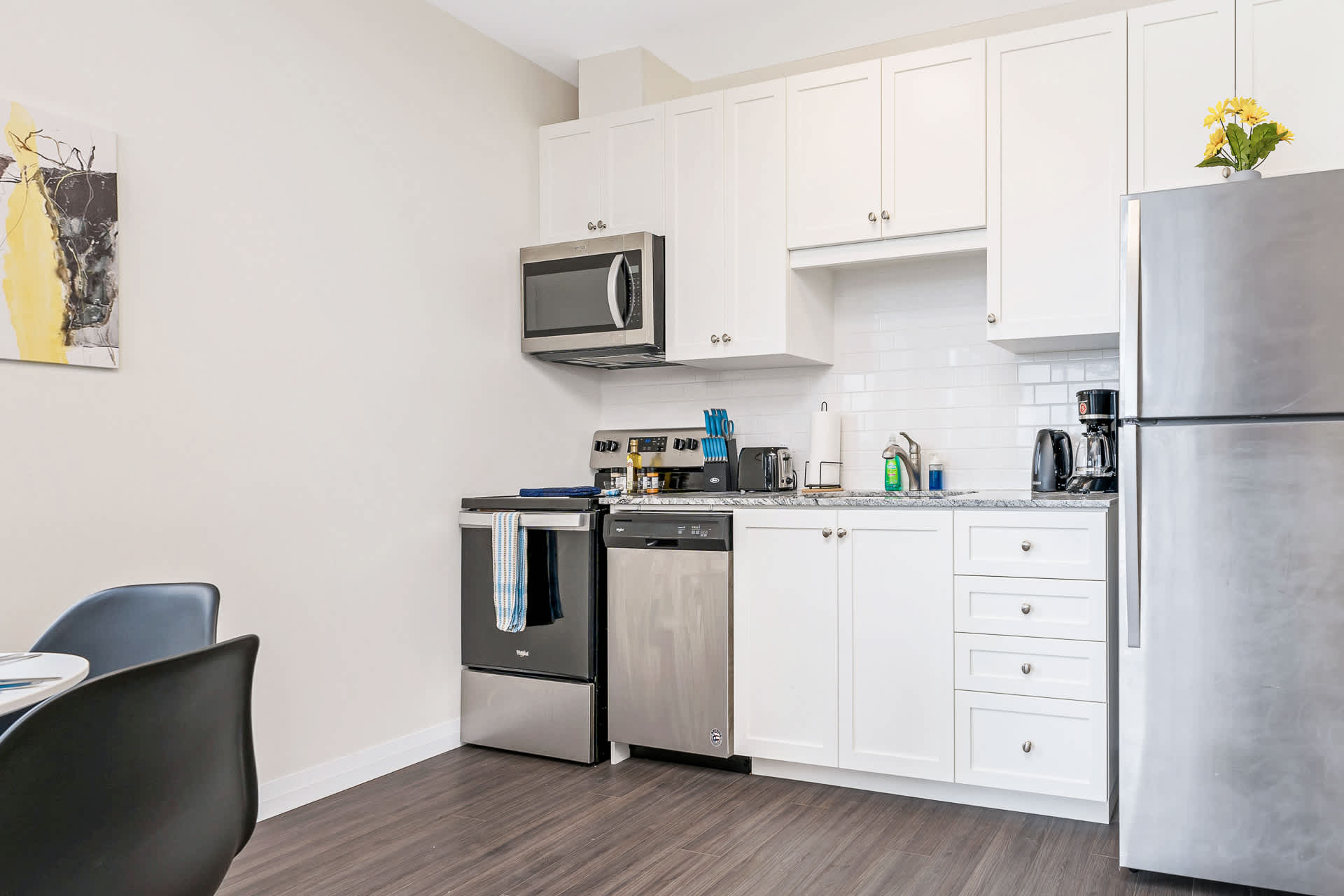 Modern kitchen featuring white shaker-style cabinets, granite countertops, and stainless steel appliances including a microwave, stove, dishwasher, and refrigerator. Dark wood-look flooring extends into a small dining area.
