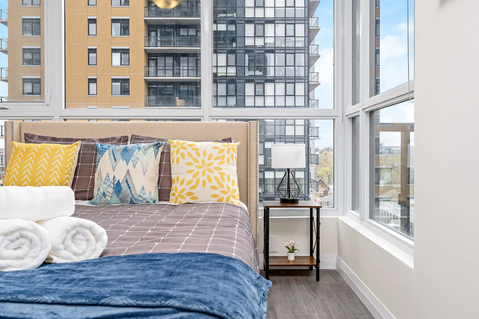 Bright bedroom with a queen-sized bed featuring a beige upholstered headboard and patterned pillows. A modern nightstand with a geometric lamp sits beside large windows offering a city view of high-rise buildings.