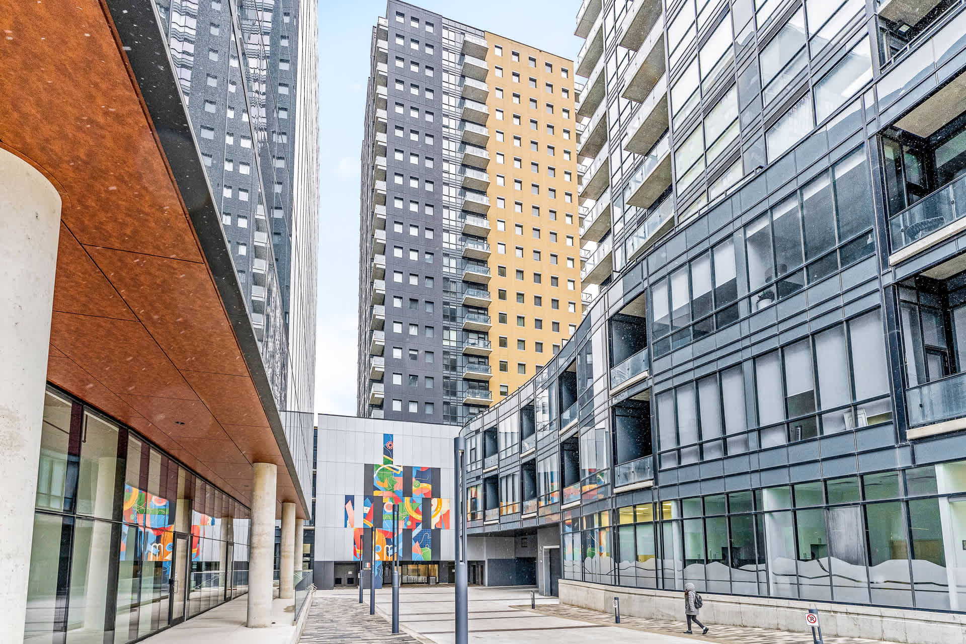 Exterior view of modern high-rise buildings with varied architectural designs, including a building with yellow and black panels. A paved courtyard with streetlights and a colorful mural is visible at the base.