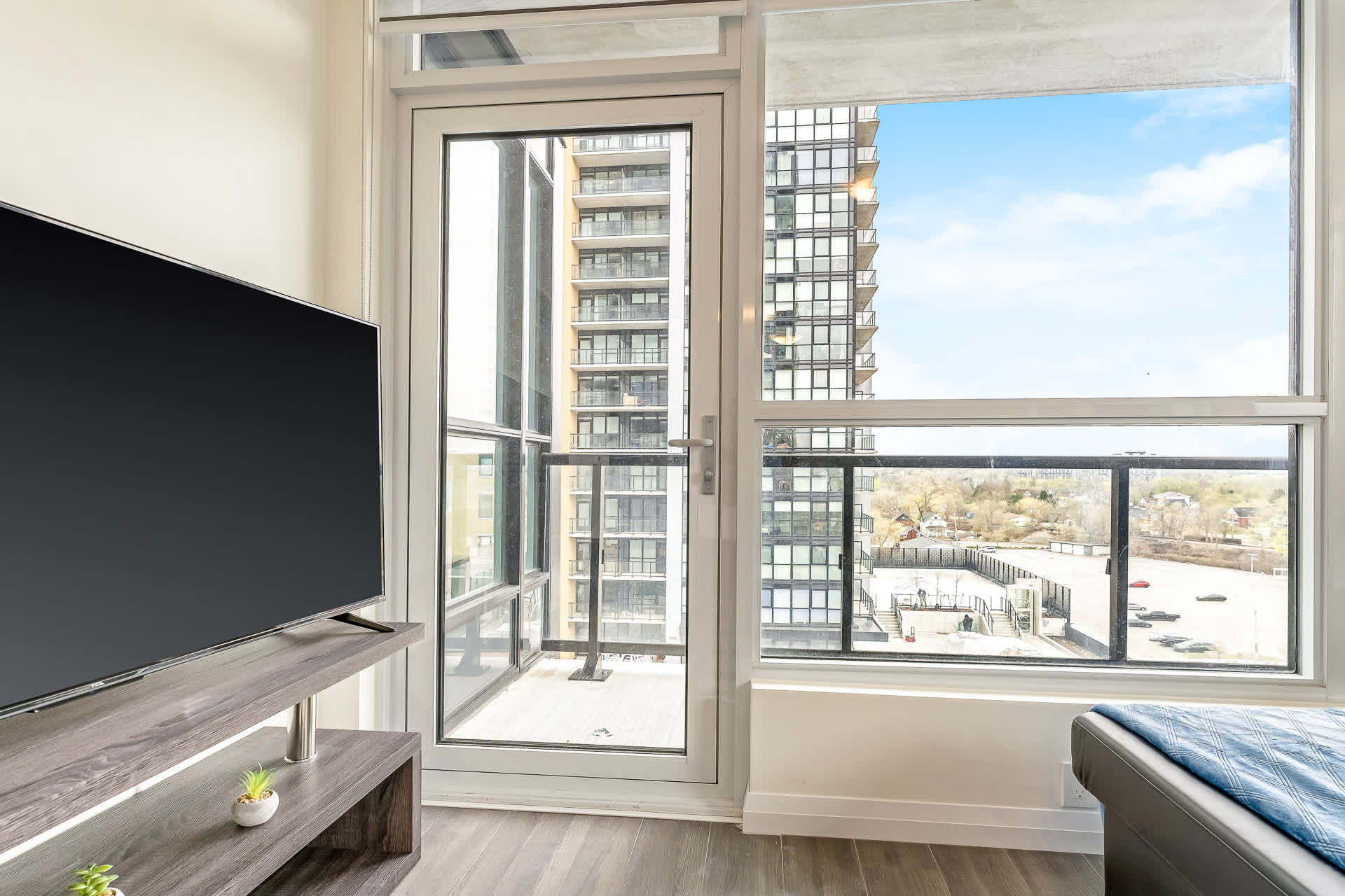 Close-up view of the living room's large windows and balcony door, showcasing the high-rise building exterior and distant city landscape. A flat-screen TV on a dark wood stand is visible on the left.