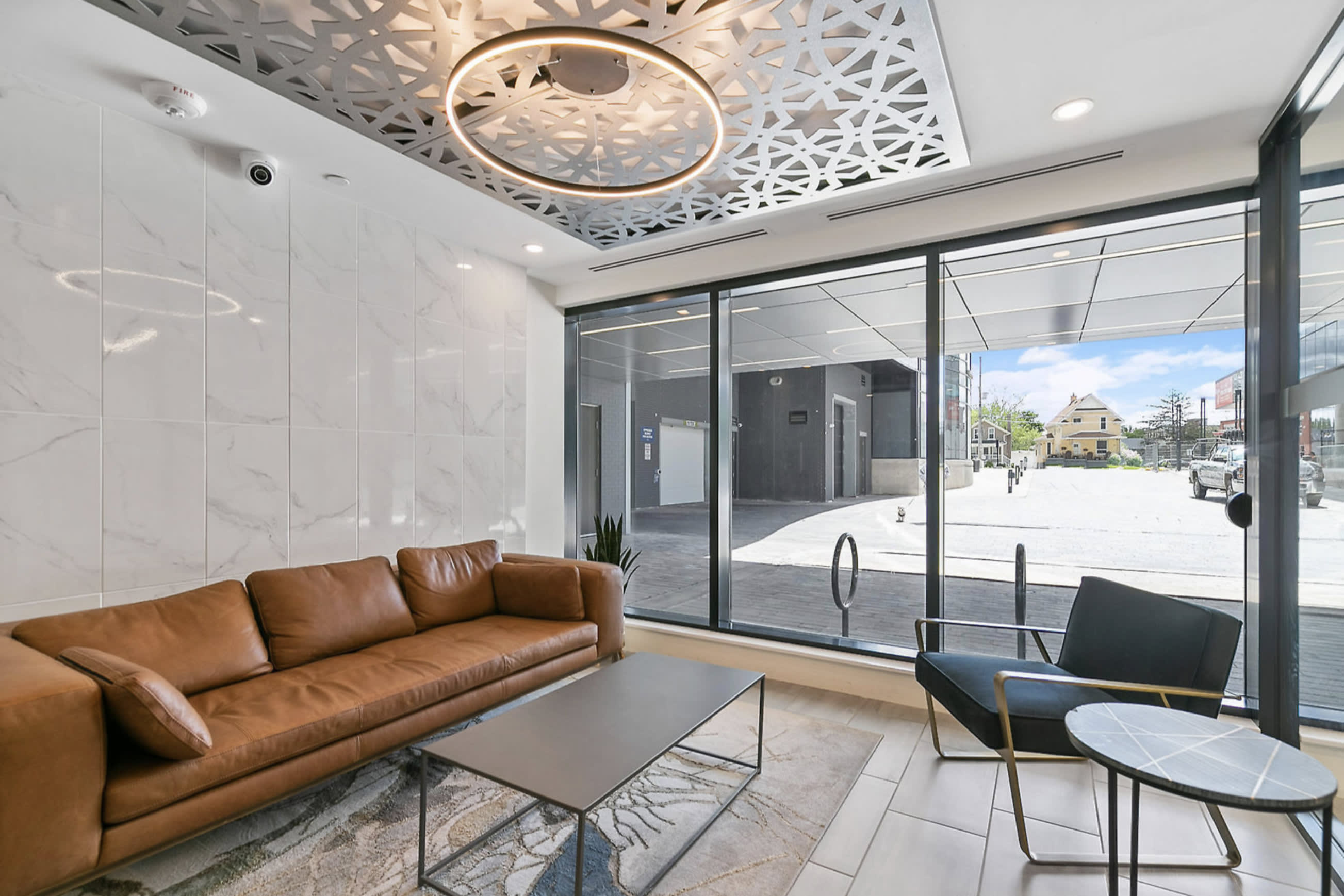 Stylish common area or lobby featuring a long brown leather sofa, a modern coffee table, and an accent chair. Large windows overlook the exterior, and the ceiling has an intricate geometric design with a circular light fixture.