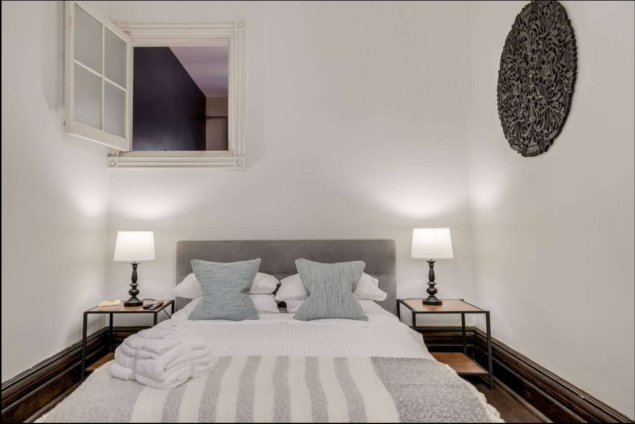 This cozy bedroom features a grey upholstered queen bed with light blue pillows, flanked by dark wood nightstands with matching lamps. A unique open window frame above the bed adds architectural interest, complemented by a dark circular wall art piece.