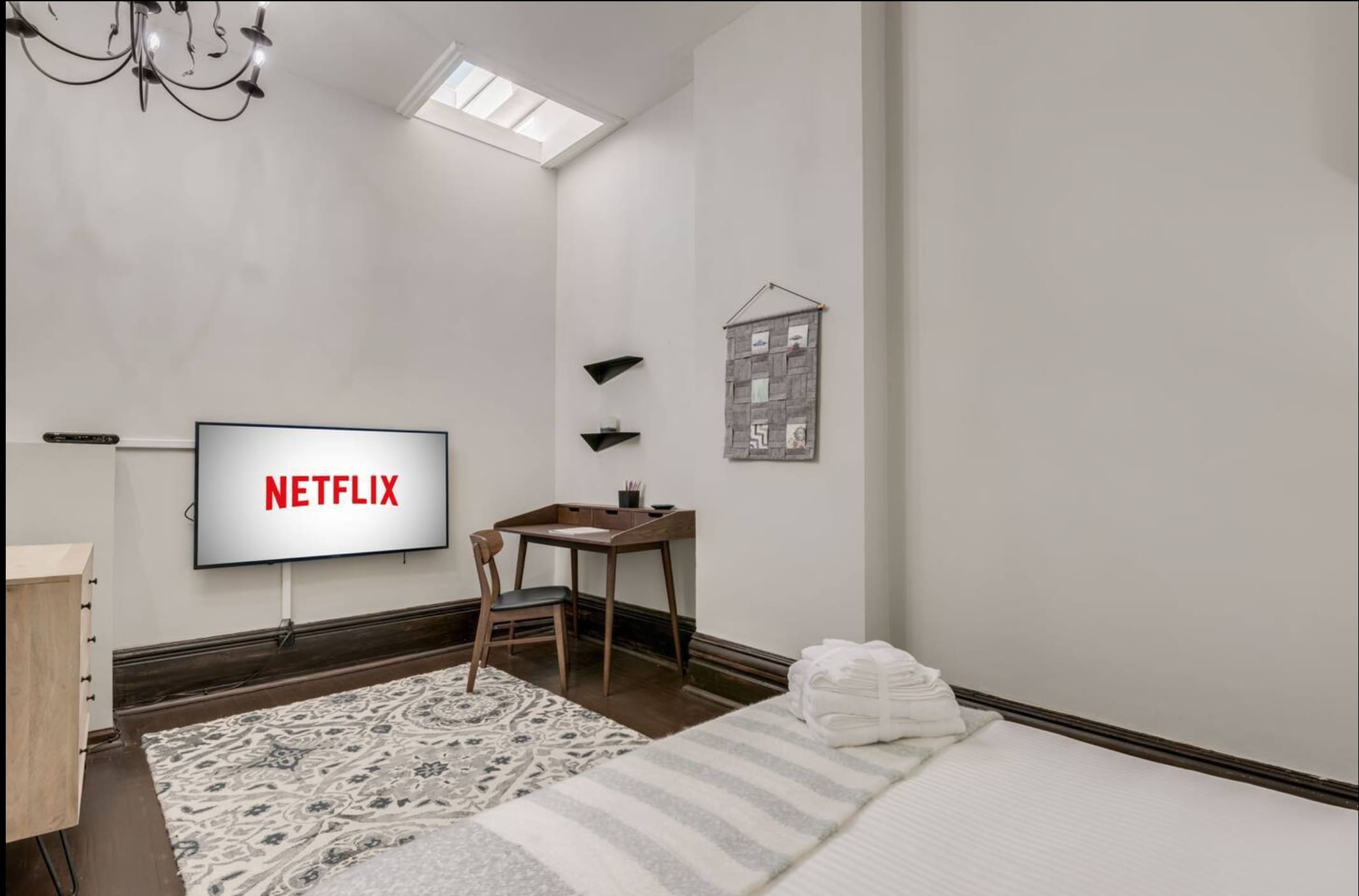 A modern bedroom with a large wall-mounted TV displaying Netflix, a minimalist wooden desk with a chair, and floating shelves. A patterned rug covers part of the dark hardwood floor, and a skylight brightens the space.