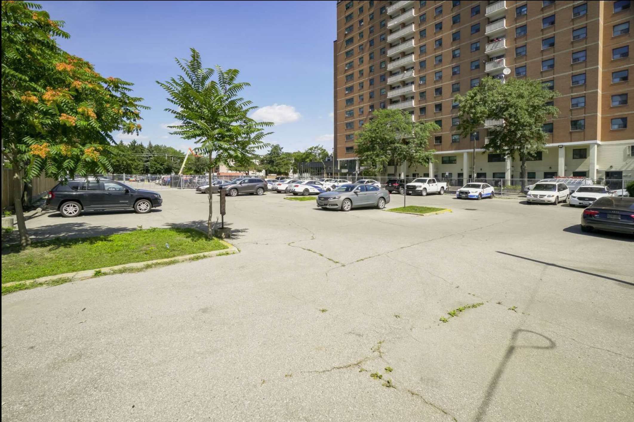 A spacious outdoor parking lot with numerous cars, surrounded by trees and a large multi-story apartment building in the background under a clear blue sky.