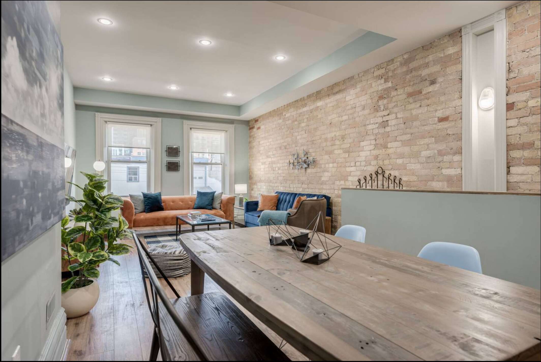 View of the open concept dining area with a rustic wooden table and light blue chairs, looking towards the living room. A large potted plant adds greenery, and light wood floors connect the spaces.