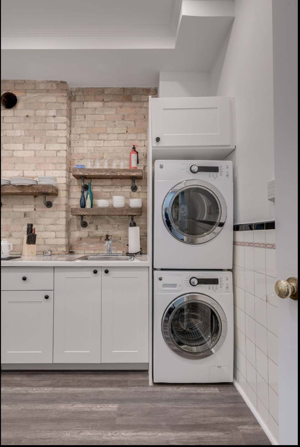 This compact kitchen features white shaker cabinets, a sink, and rustic wooden shelves against an exposed brick wall. A stacked washer and dryer unit is neatly integrated into the space, providing laundry facilities.