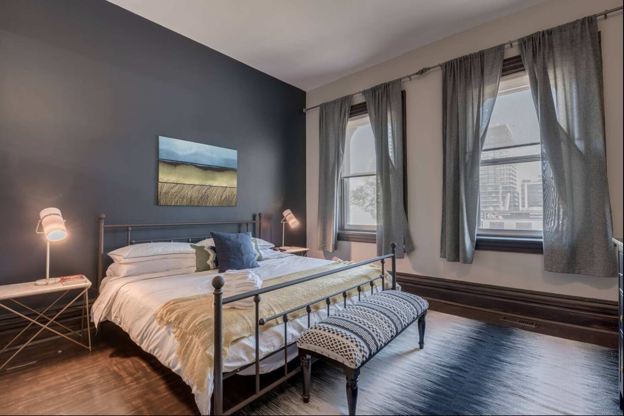 A bedroom with a dark blue accent wall behind a metal-frame bed dressed in white and yellow linens. Two large windows with grey curtains offer city views, and a patterned bench sits at the foot of the bed.