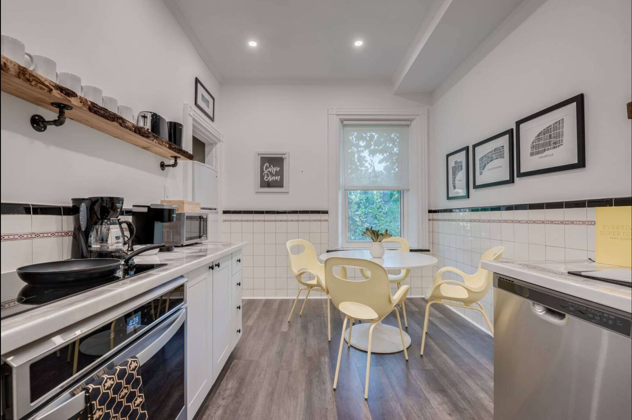 A bright dining area with a round white table and four yellow chairs, set against a white tiled wall. The kitchen counter with appliances is visible on the left, and a window with a roller shade overlooks greenery.