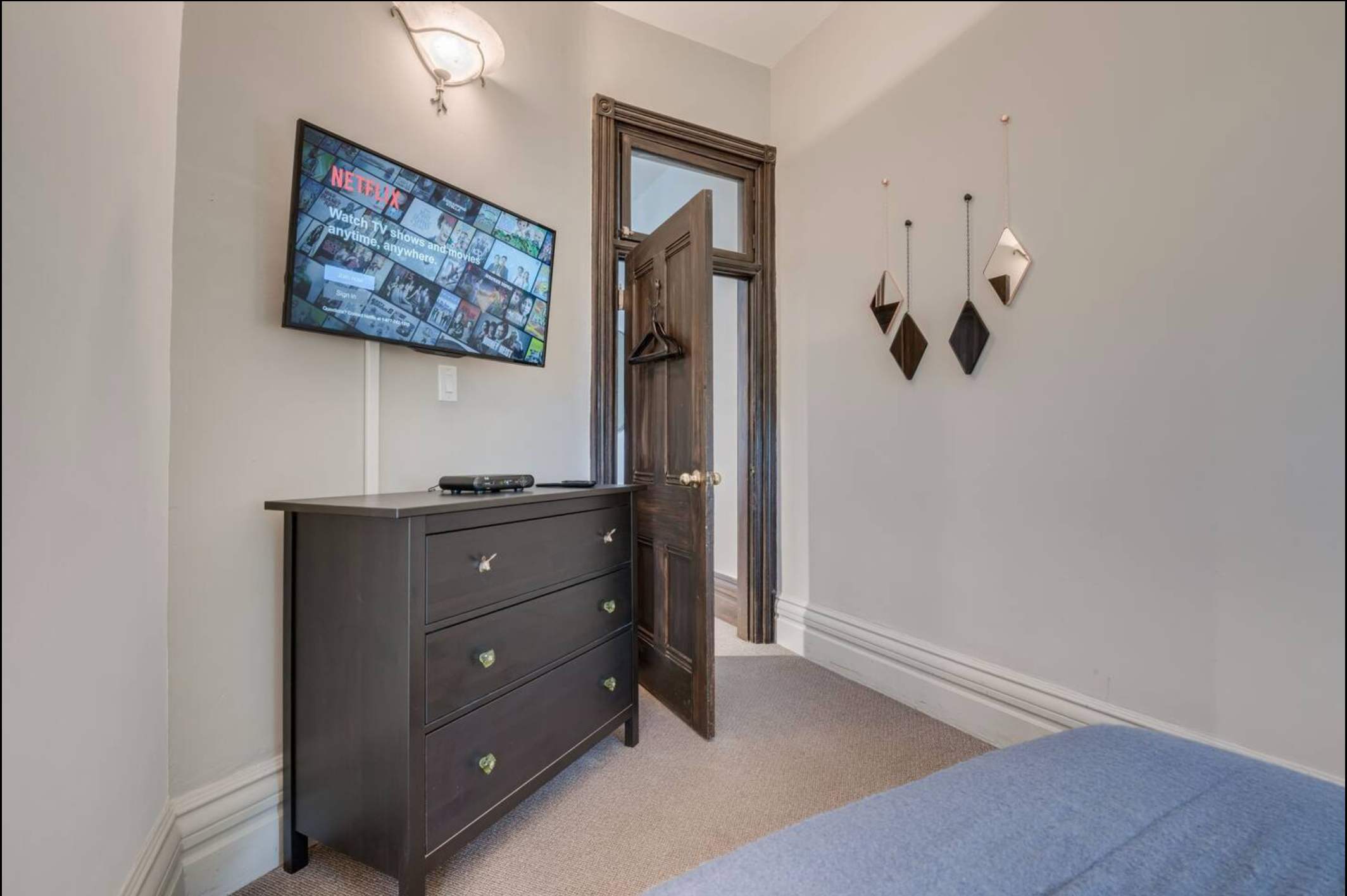 Another angle of the bedroom showing a wall-mounted flat-screen TV above a dark dresser. The rustic wooden door to the bathroom is open, revealing a toilet inside.