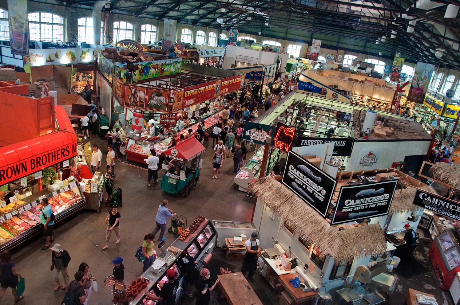 A bustling indoor market hall filled with various food stalls, vendors, and shoppers. The high ceiling with exposed beams and large windows suggests a historic or industrial building.