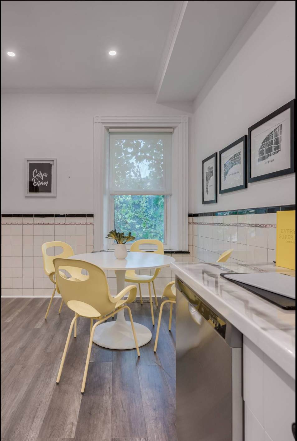 A close-up of the dining area featuring a white round table with a small plant and four modern yellow chairs. The room has white tiled walls, dark wood-look flooring, and a window with a roller shade.