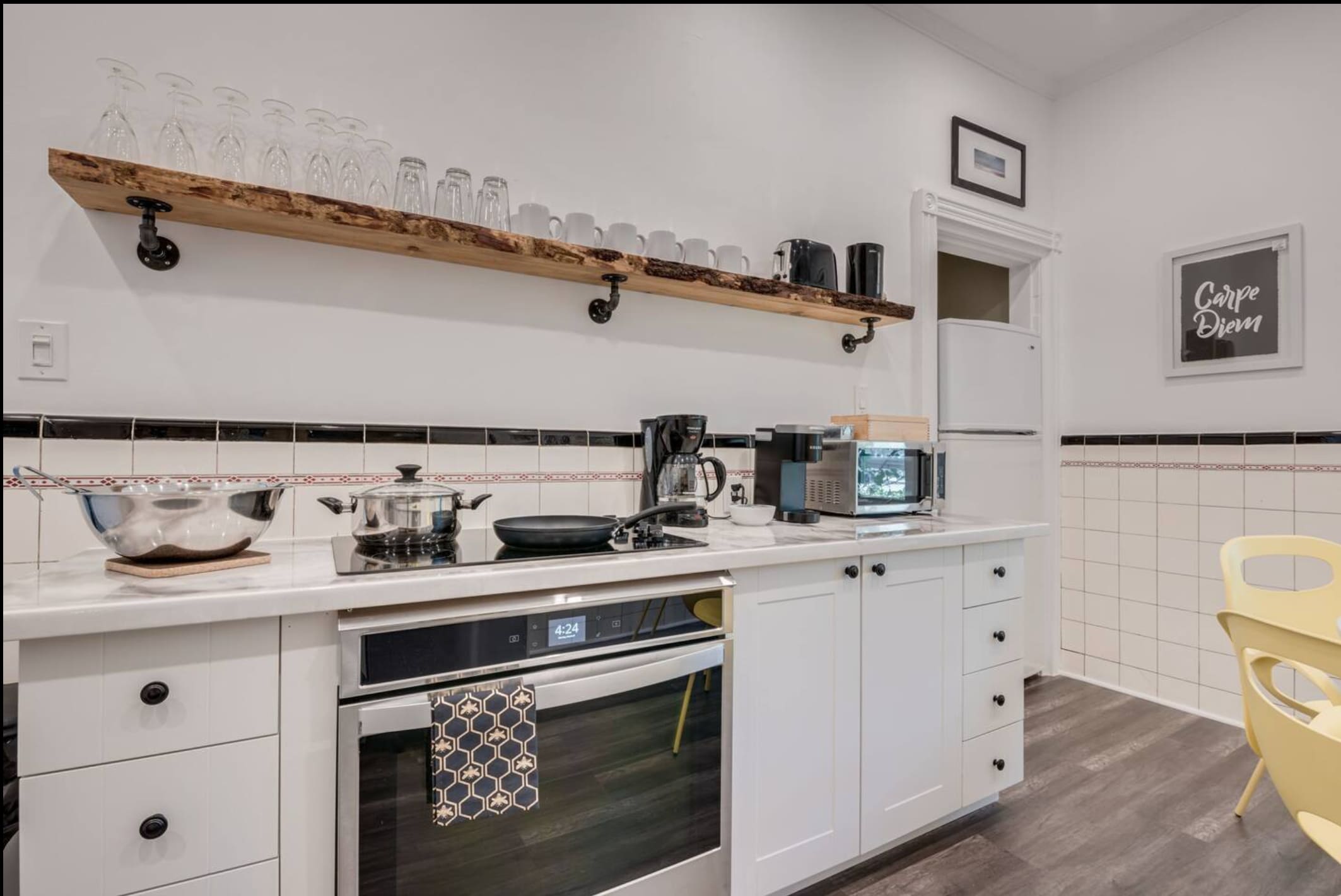 A modern kitchen with white cabinets, a white countertop, and a black and white tiled backsplash. It features a built-in oven, electric stovetop, coffee maker, and a rustic wooden shelf with glassware.