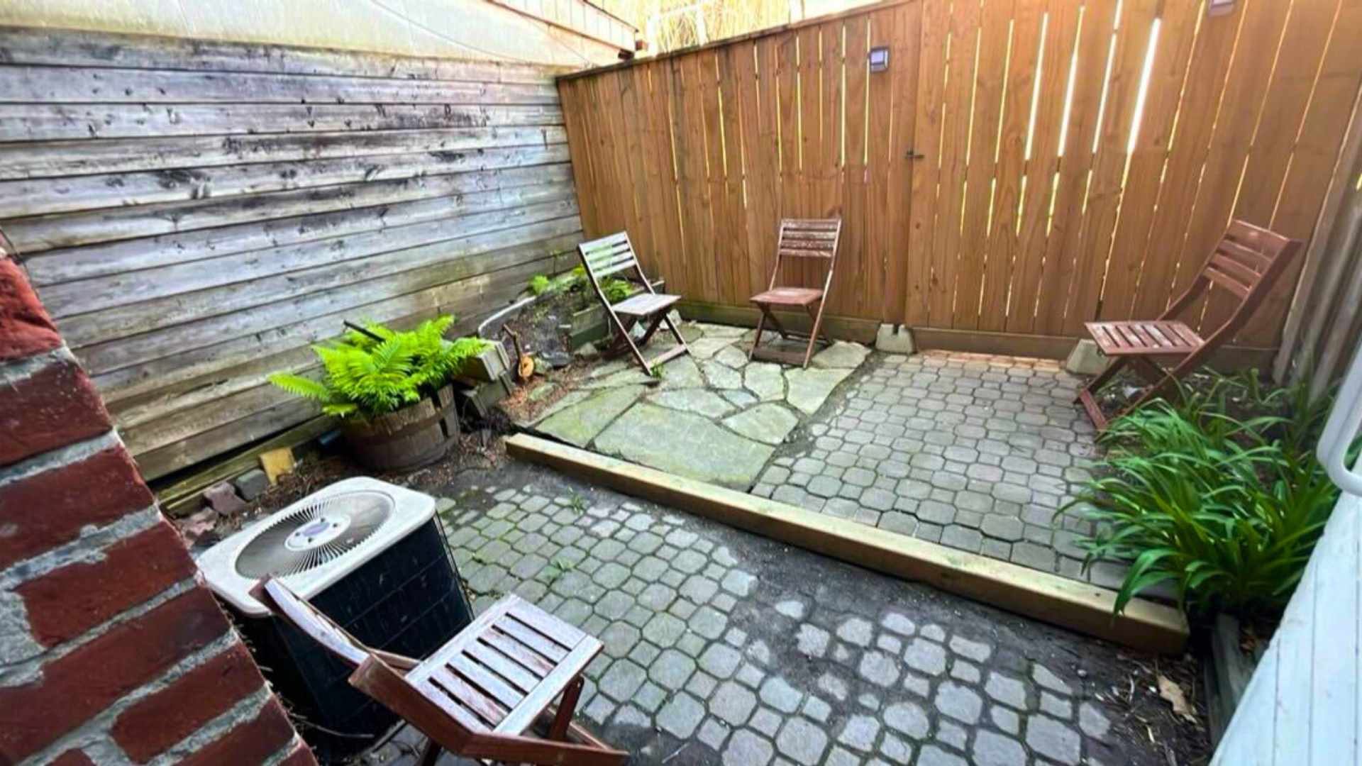A private outdoor patio with mixed stone and paver flooring, enclosed by wooden fences. Several rustic wooden chairs are arranged around the space, with an air conditioning unit visible in the foreground.