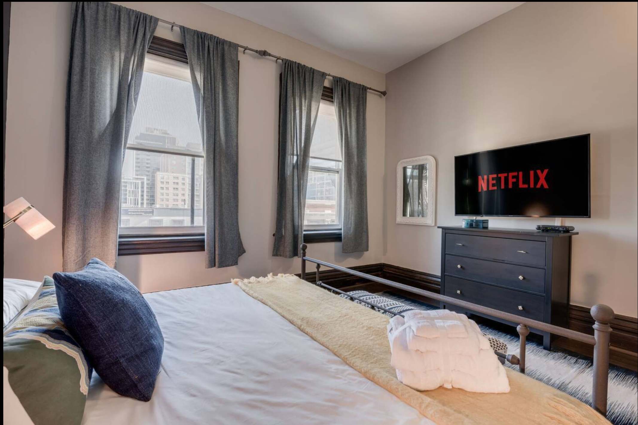 Another view of the bedroom showing a large flat-screen TV on a dark dresser opposite the metal-frame bed. Three tall windows with grey curtains provide natural light and city views.