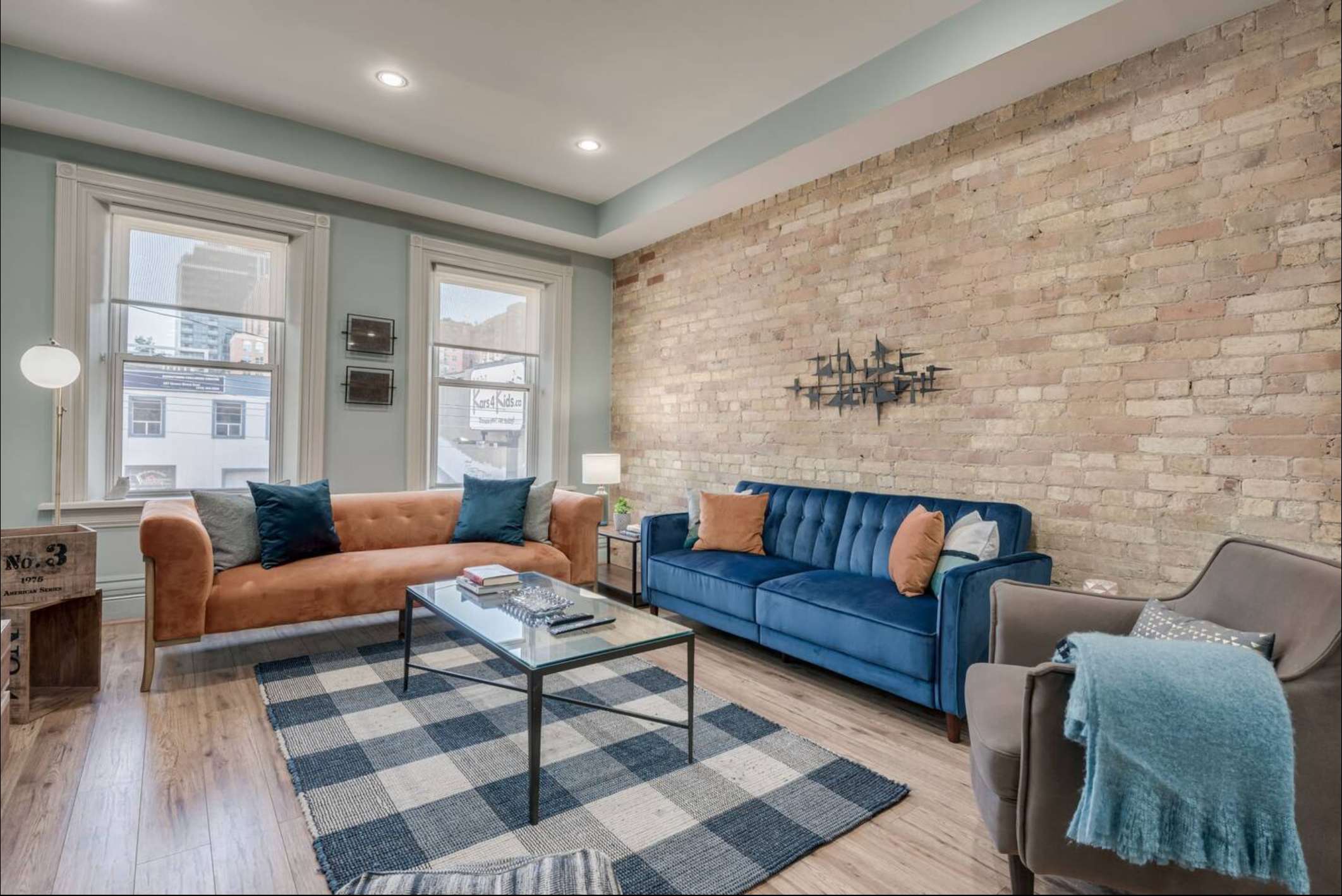 Modern living room with light blue walls and an exposed brick accent wall. Features a burnt orange sofa, a blue sofa, a gray armchair, and a glass coffee table on a blue and white plaid rug.