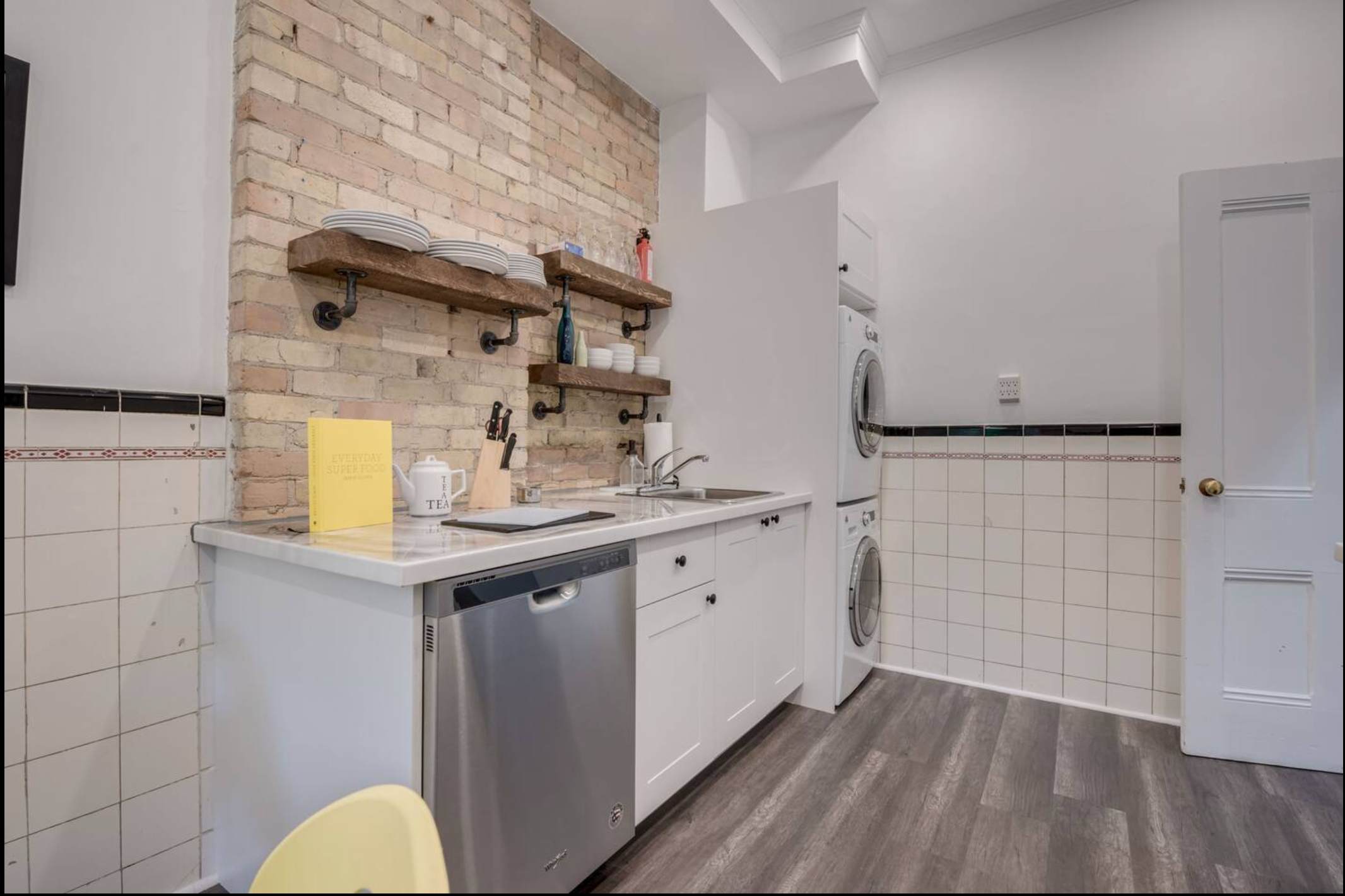A kitchen area with white cabinets, a stainless steel dishwasher, and a brick accent wall. Stacked washer and dryer units are visible next to the counter, and a white tiled wall extends around the room.