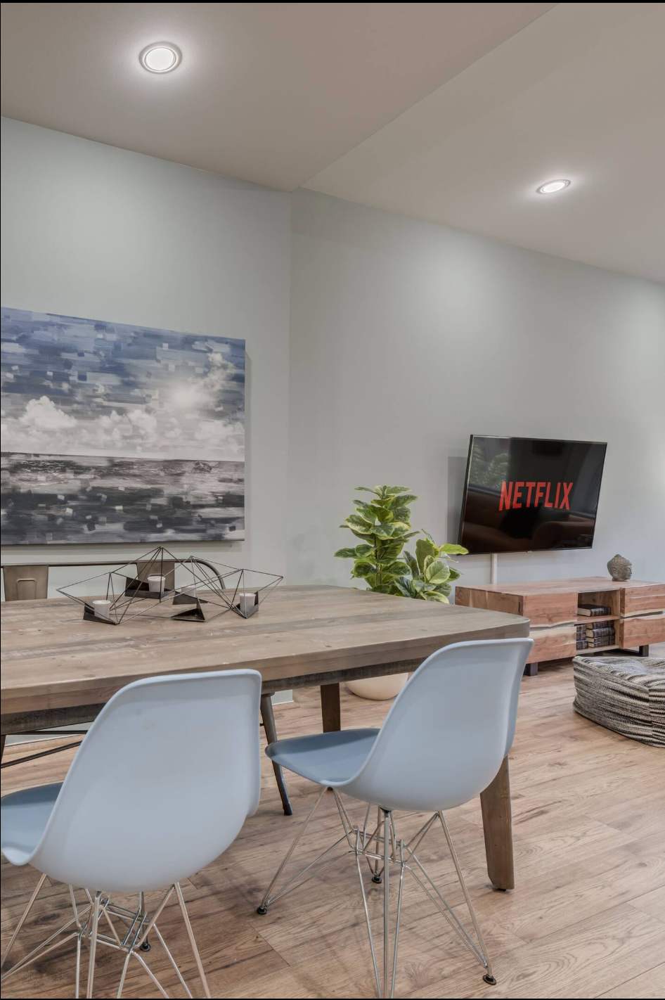 Close-up view of the dining area with a rustic wooden table and light blue chairs, showing the adjacent living room with a TV. A large abstract painting and a potted plant decorate the space.
