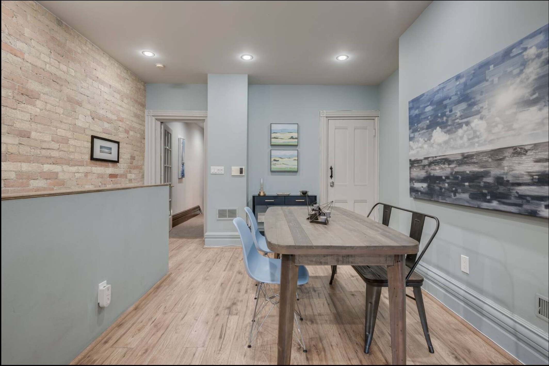 Dining area featuring a rustic wooden table with light blue and metal chairs, set against light blue walls. An abstract painting hangs above the table, and light wood floors lead to a hallway with a door.