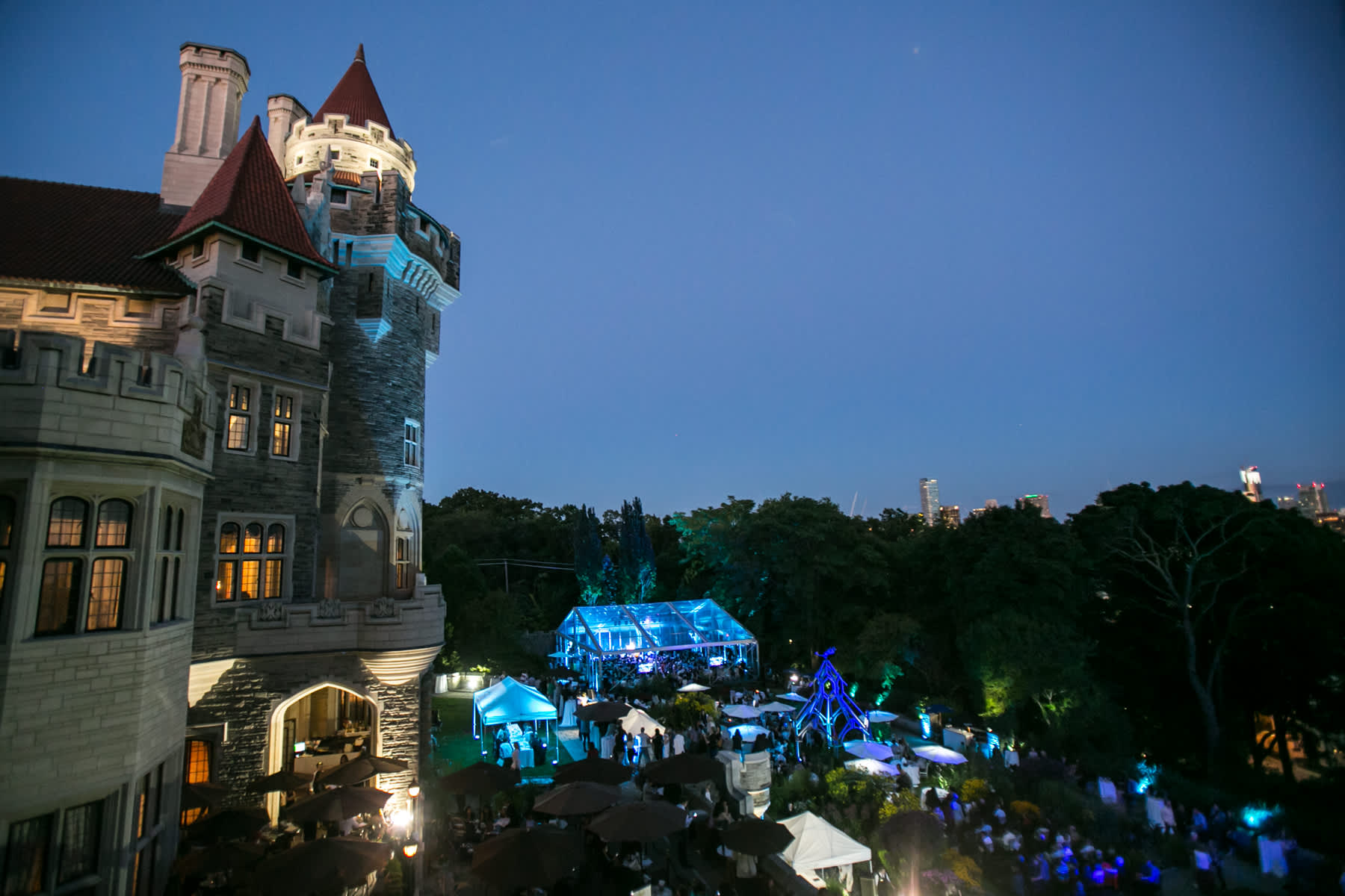 An aerial view at dusk of a grand castle-like building with turrets and an outdoor event space illuminated with blue lights. Numerous people are gathered under tents and umbrellas, with a city skyline visible in the distance.