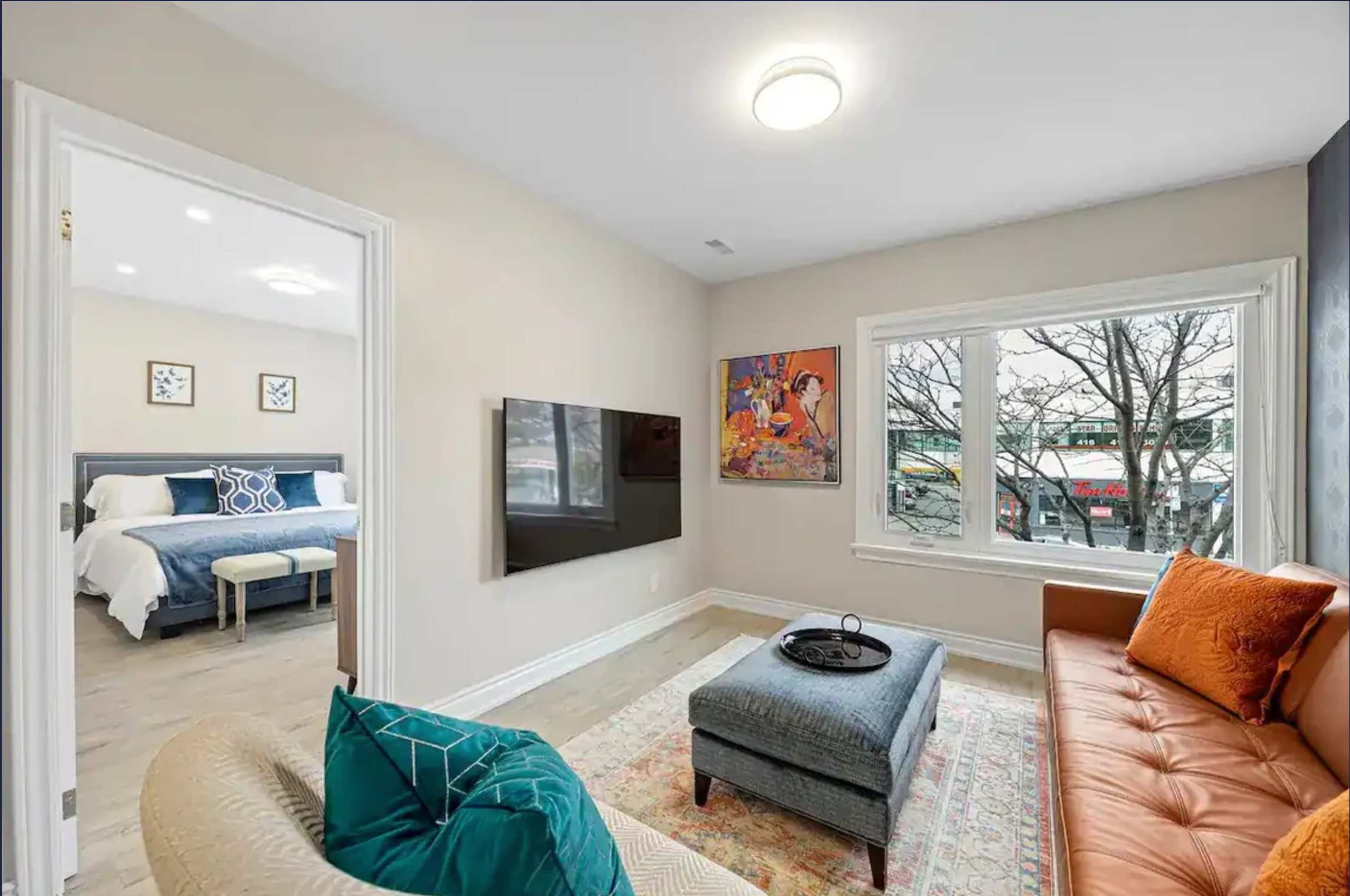 A cozy living room with a tan leather sofa, a cream armchair, a grey ottoman serving as a coffee table, and a wall-mounted TV, with a bedroom visible through an open doorway.