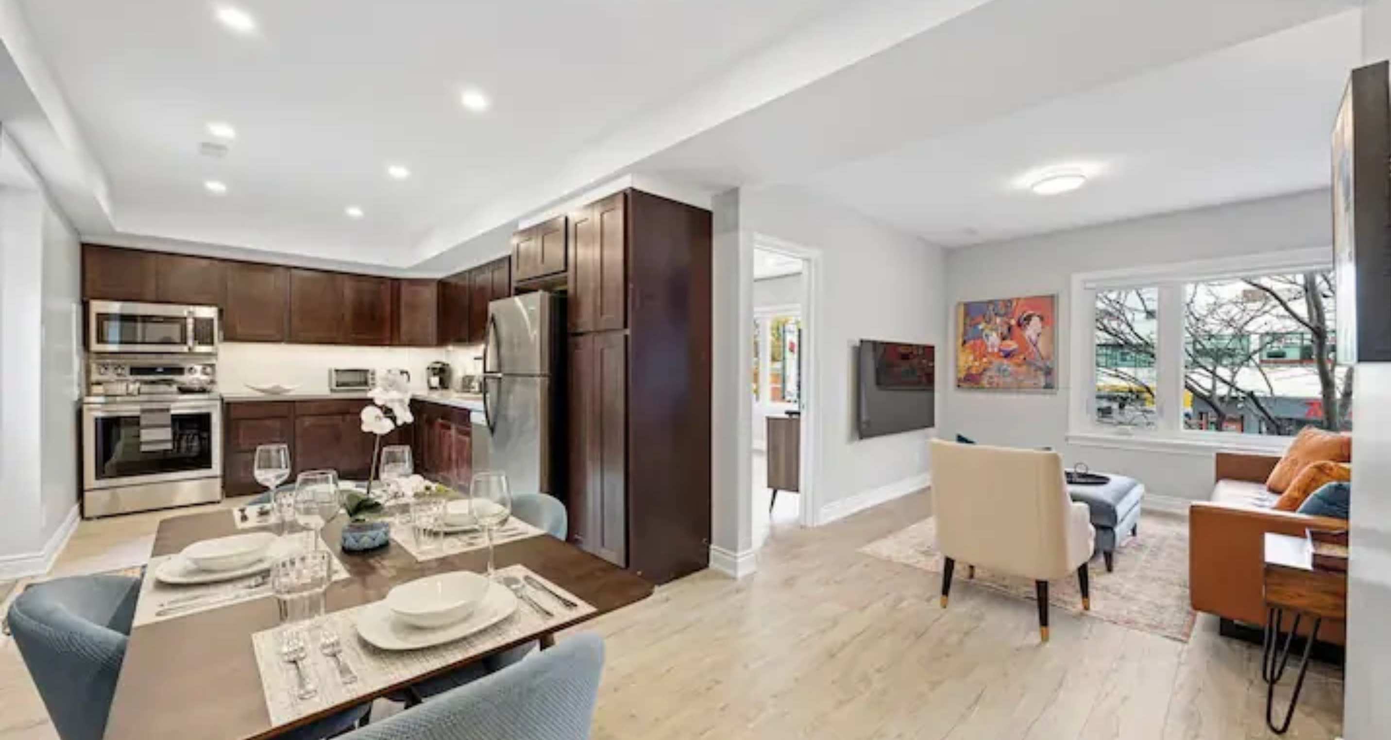 An open-concept space featuring a dark wood dining table set for four, a fully equipped kitchen with dark cabinetry and stainless steel appliances, and a glimpse of the living room with a sofa and window.