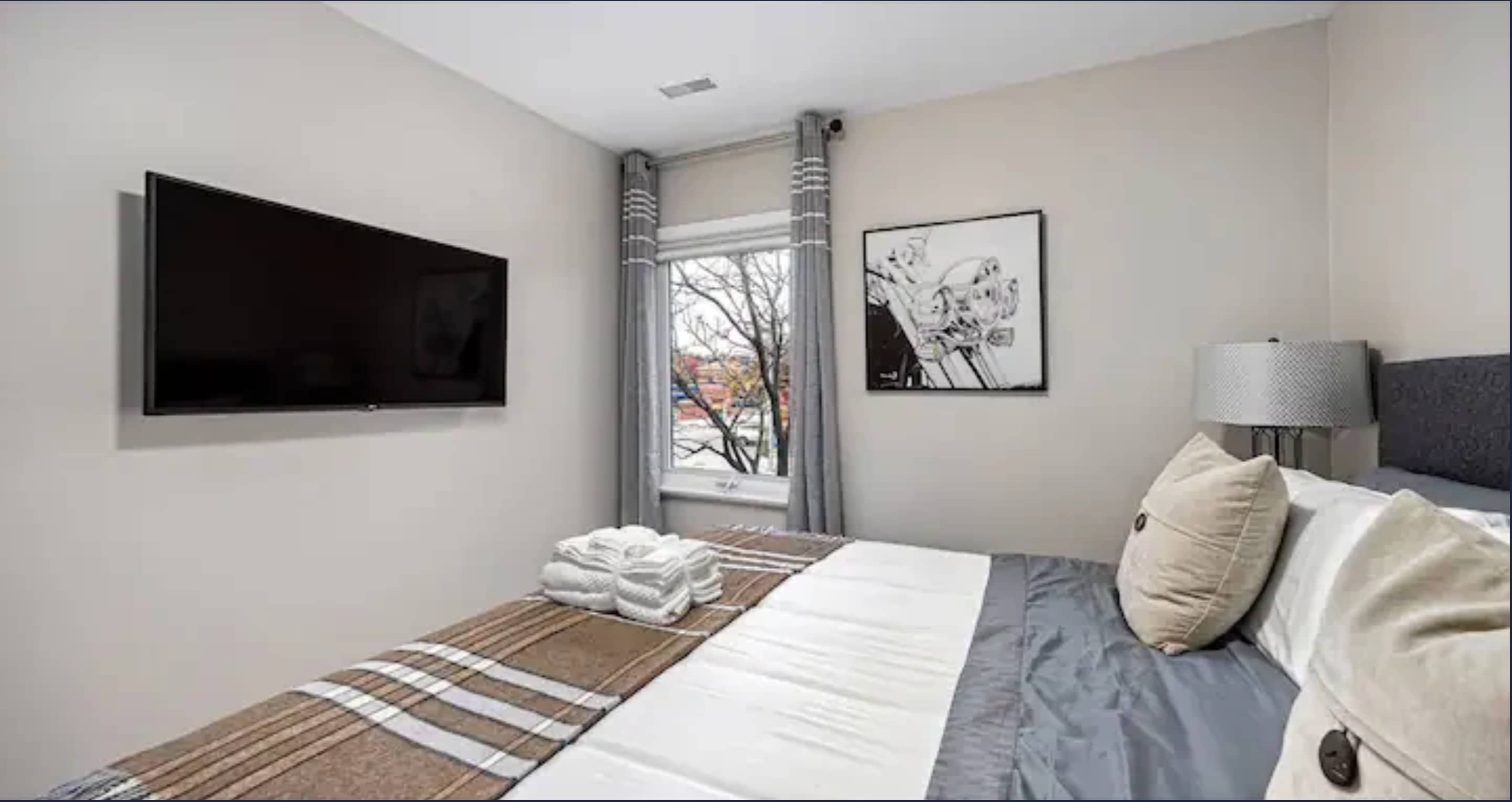 View of the second bedroom showing a wall-mounted flat-screen TV opposite the queen-sized bed with grey and brown plaid bedding. A window with grey curtains allows natural light, and a framed artwork hangs on the light-colored wall.