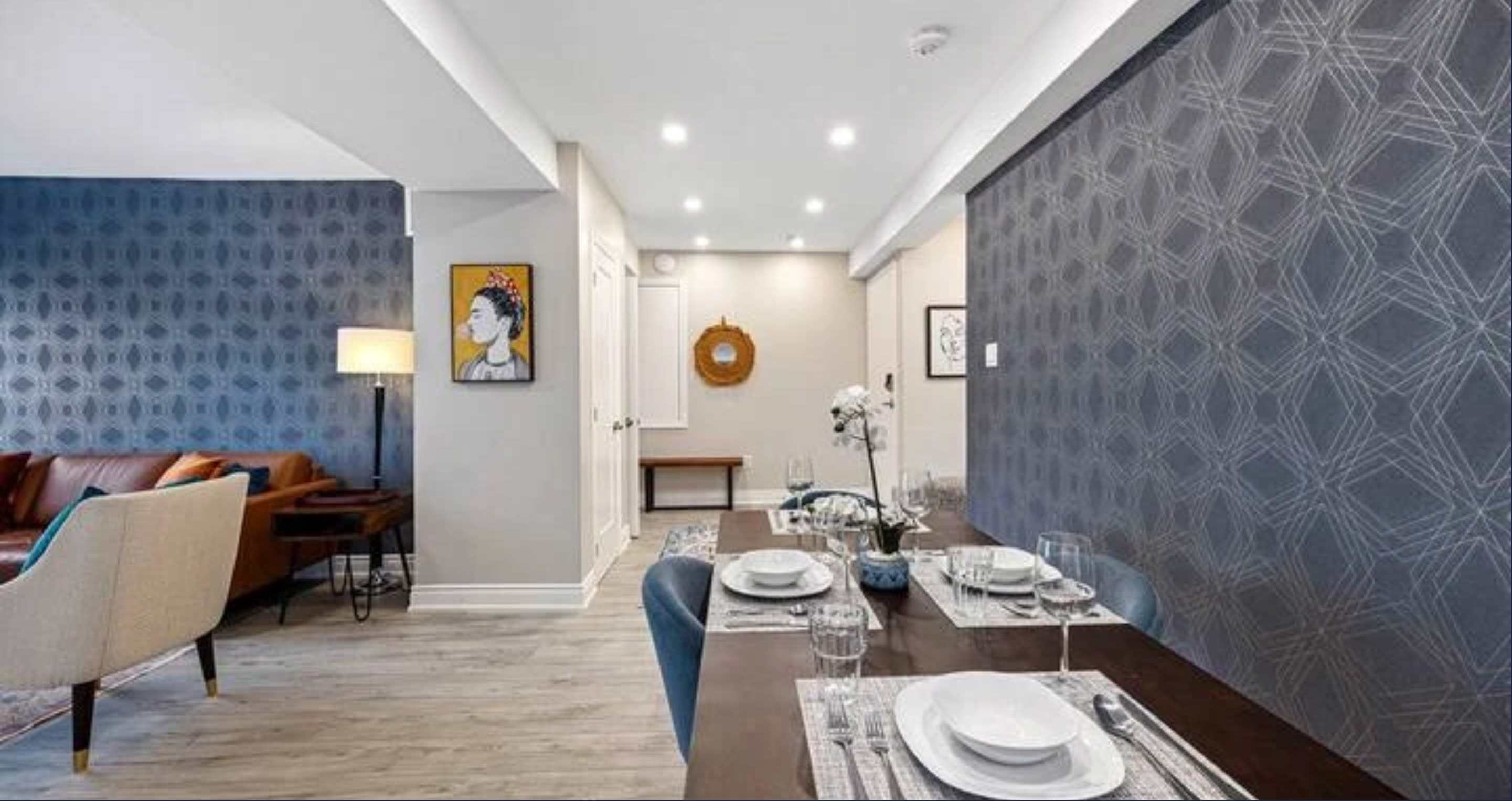 A long view of the dining area with a dark wood table and blue chairs, flanked by a patterned accent wall, leading to a small foyer area with artwork and a bench.