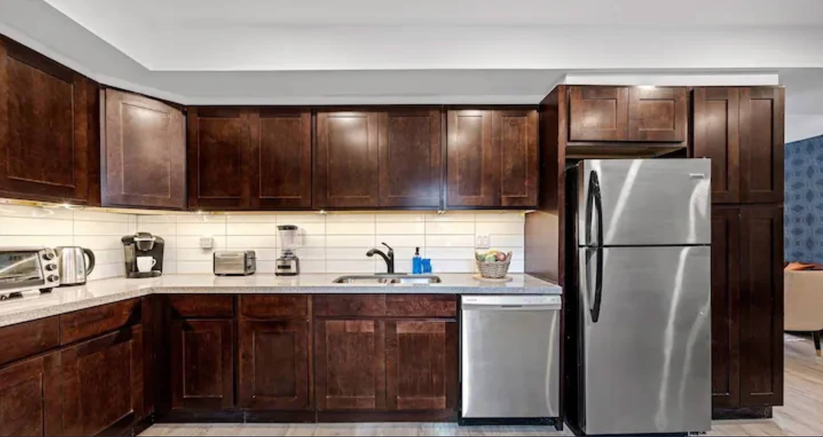 A close-up view of the kitchen showing dark wood upper and lower cabinets, light countertops, a white subway tile backsplash, and various small appliances on the counter.