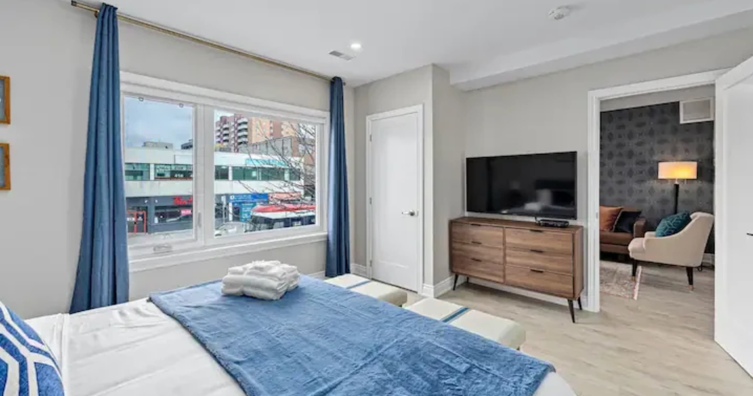 Bright bedroom with a queen-sized bed featuring blue and white bedding, a large window offering city views, and a wall-mounted TV above a wooden dresser. Blue curtains frame the window, and a doorway leads to an adjacent room.