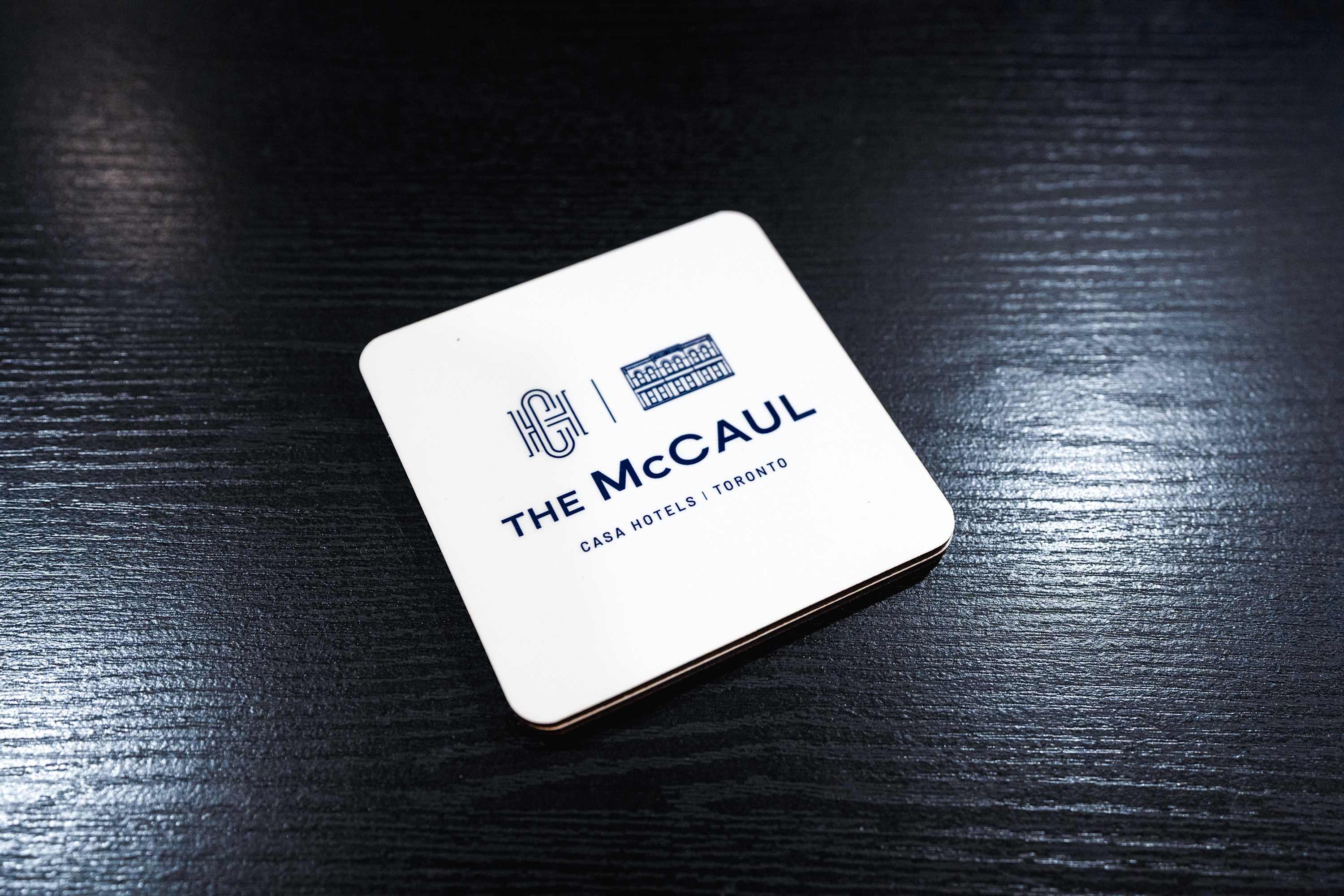 A white square coaster with "THE McCAUL | CASA HOTELS | TORONTO" printed in blue, resting on a dark, textured surface.