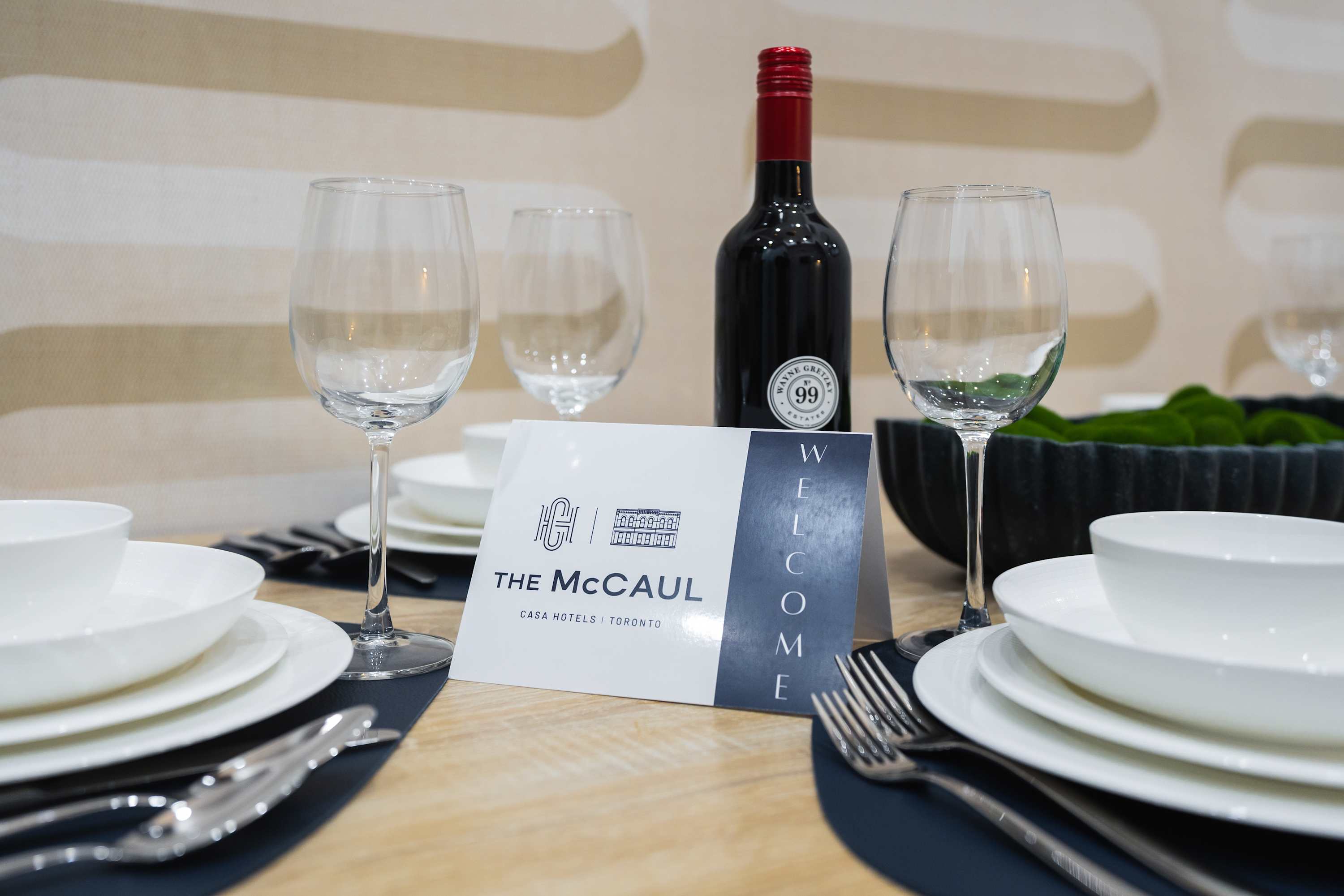A modern dining table set for four with white dinnerware, wine glasses, and a bottle of red wine, featuring a 'The McCaul Welcome' card on a light wood surface.