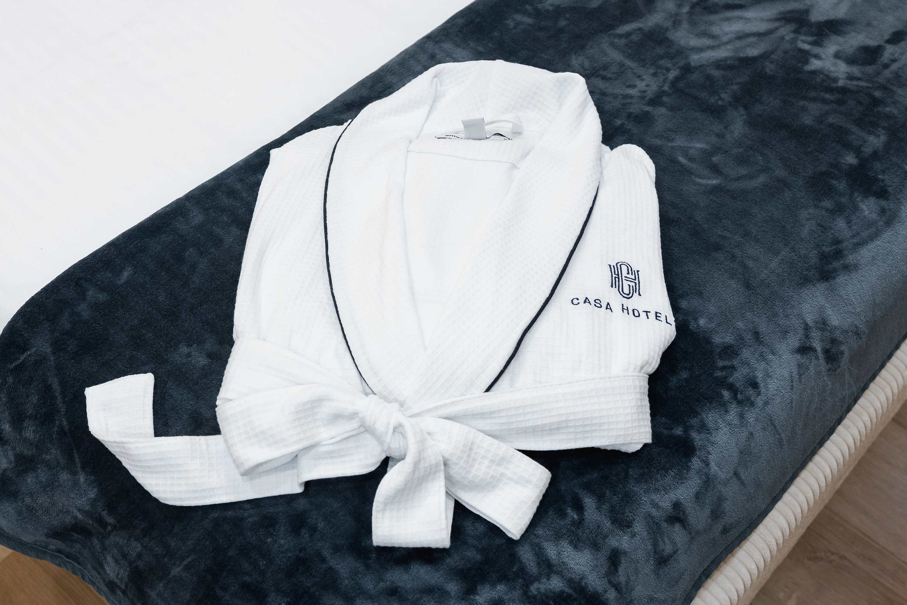 A white waffle-knit bathrobe with a dark blue trim and 'CASA HOTEL' logo embroidered on the chest, neatly folded on a dark grey upholstered bench.
