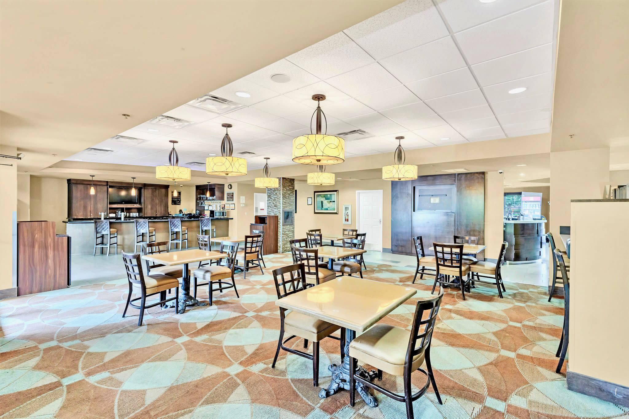 Spacious dining area with multiple square and round tables, upholstered chairs, and decorative pendant lighting, featuring a colorful patterned carpet and a bar area in the background.