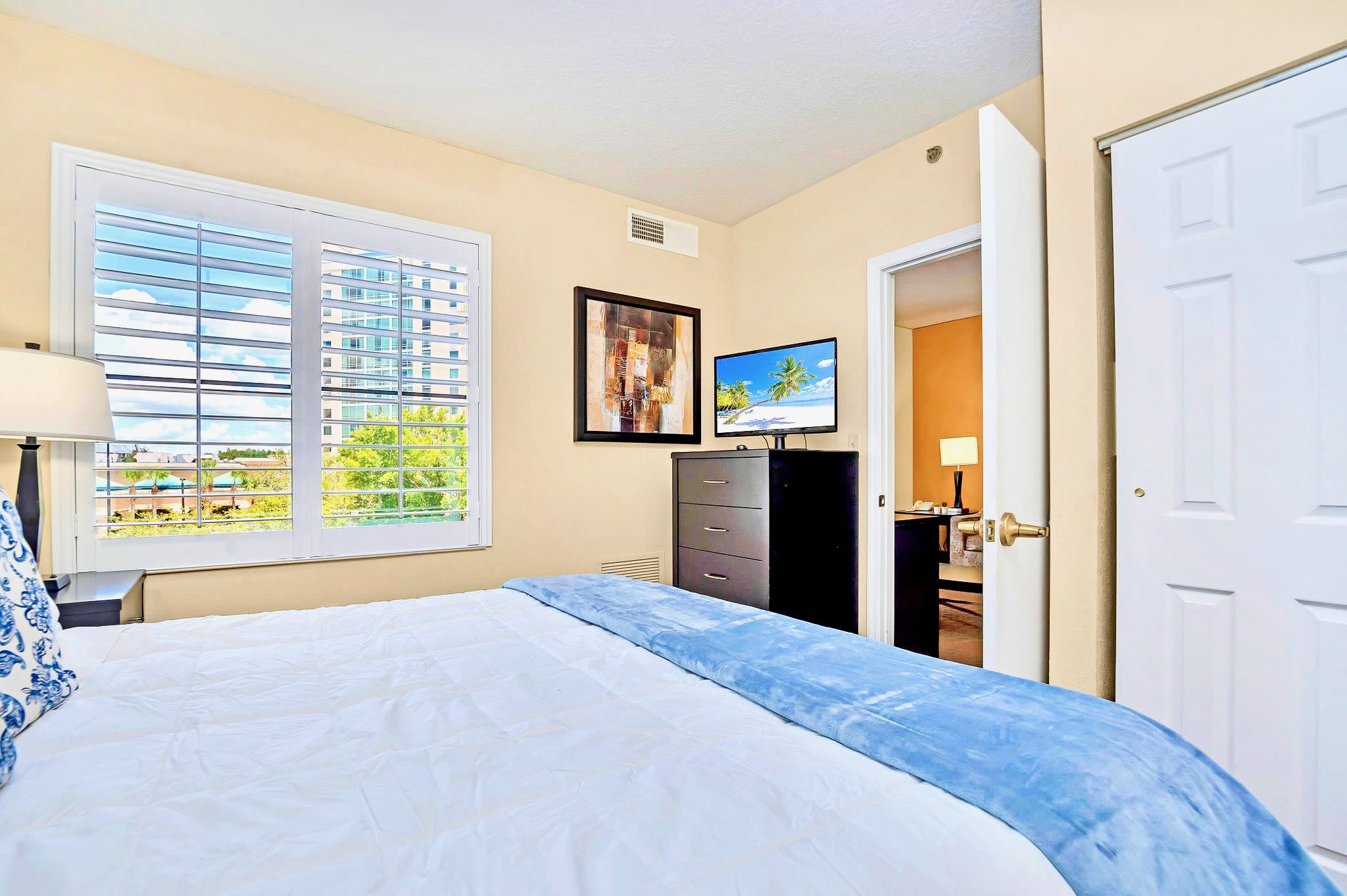 A bright bedroom features a queen-sized bed, a dark dresser with a TV, and a large window with white plantation shutters, offering a glimpse of the city outside.