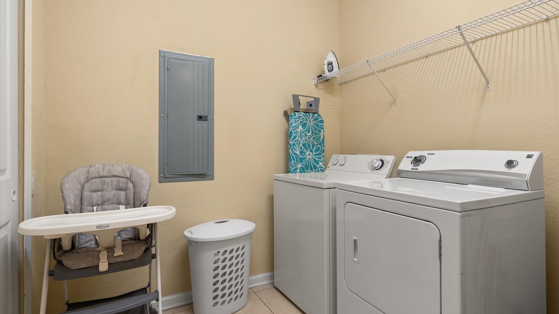 Functional laundry room with a white top-load washer and front-load dryer, a high chair, and an ironing board, set against light yellow walls with a utility panel.